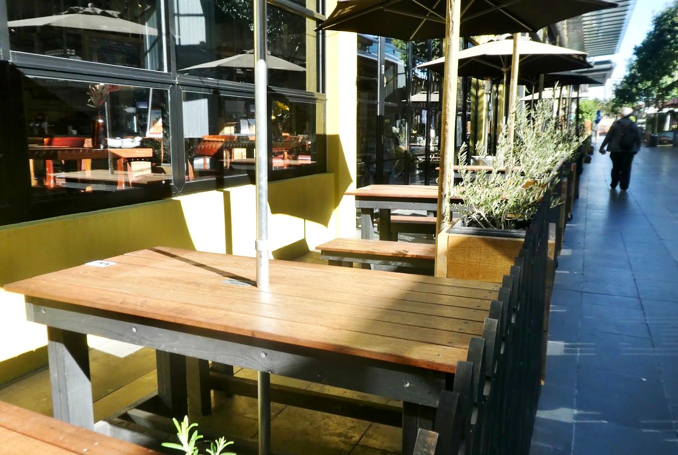 An outdoor bar area with wooden tables, potted plants, and large umbrellas along a city sidewalk at The Melbourne Cellardoor.