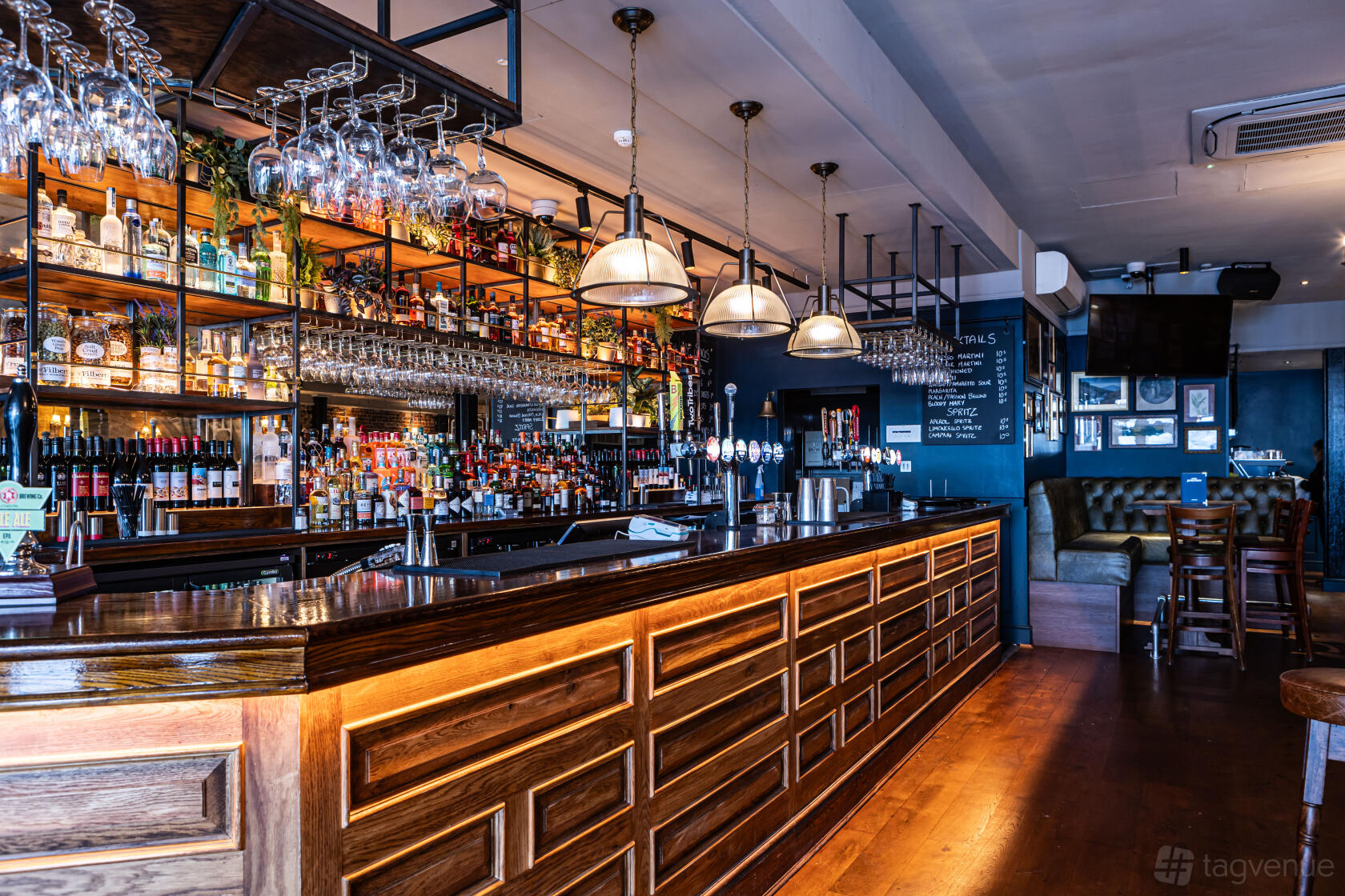 A pub with a long wooden bar, overhead glass racks, pendant lights, and leather seating at The Peacock.