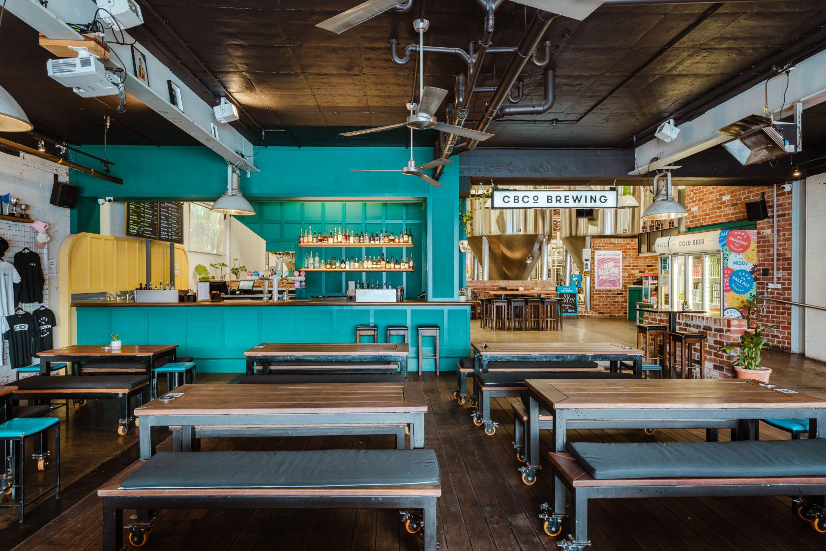 A function room with long wooden tables, bench seating, and a teal bar area at CBCo Brewing.