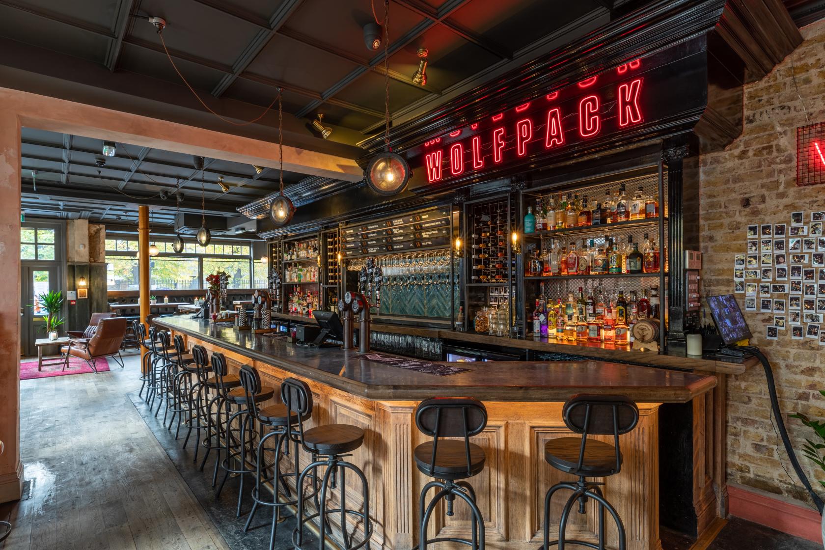 A bar with a long wooden counter, exposed brick walls, neon signage, and shelves stocked with spirits at The Wolfpack Inn.