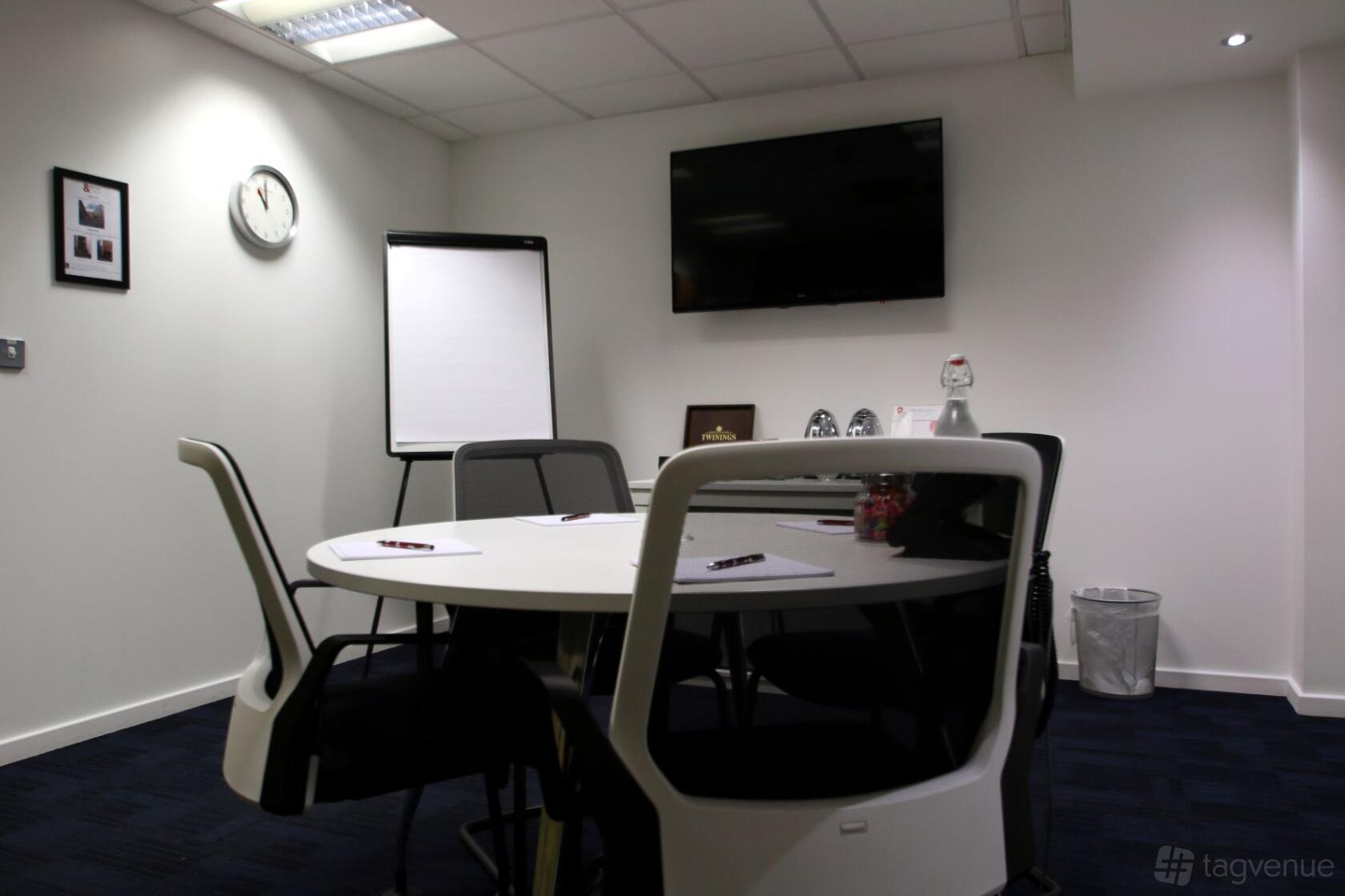 A meeting room with a round table, white chairs, wall-mounted TV, and flip chart at 150 Minories.