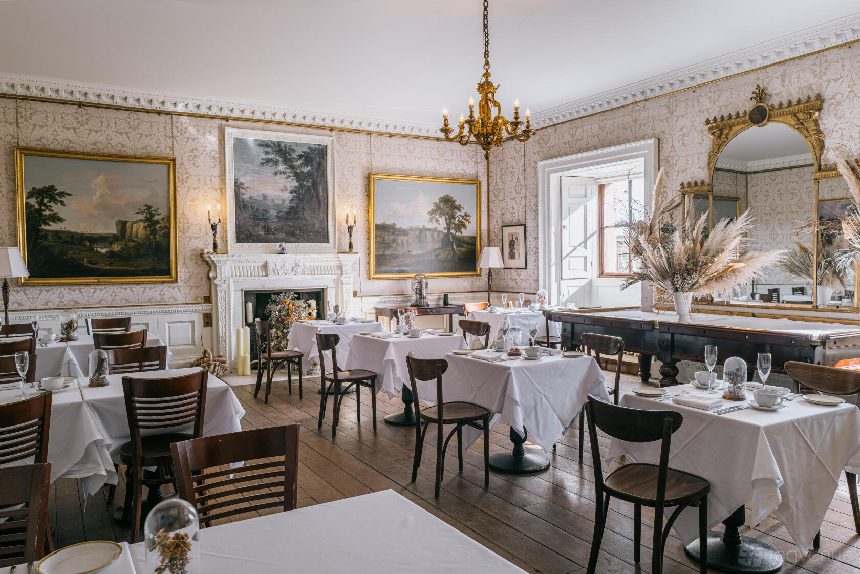 An event space with white tablecloths, wooden chairs, a chandelier, and ornate wall art at Harewood House.