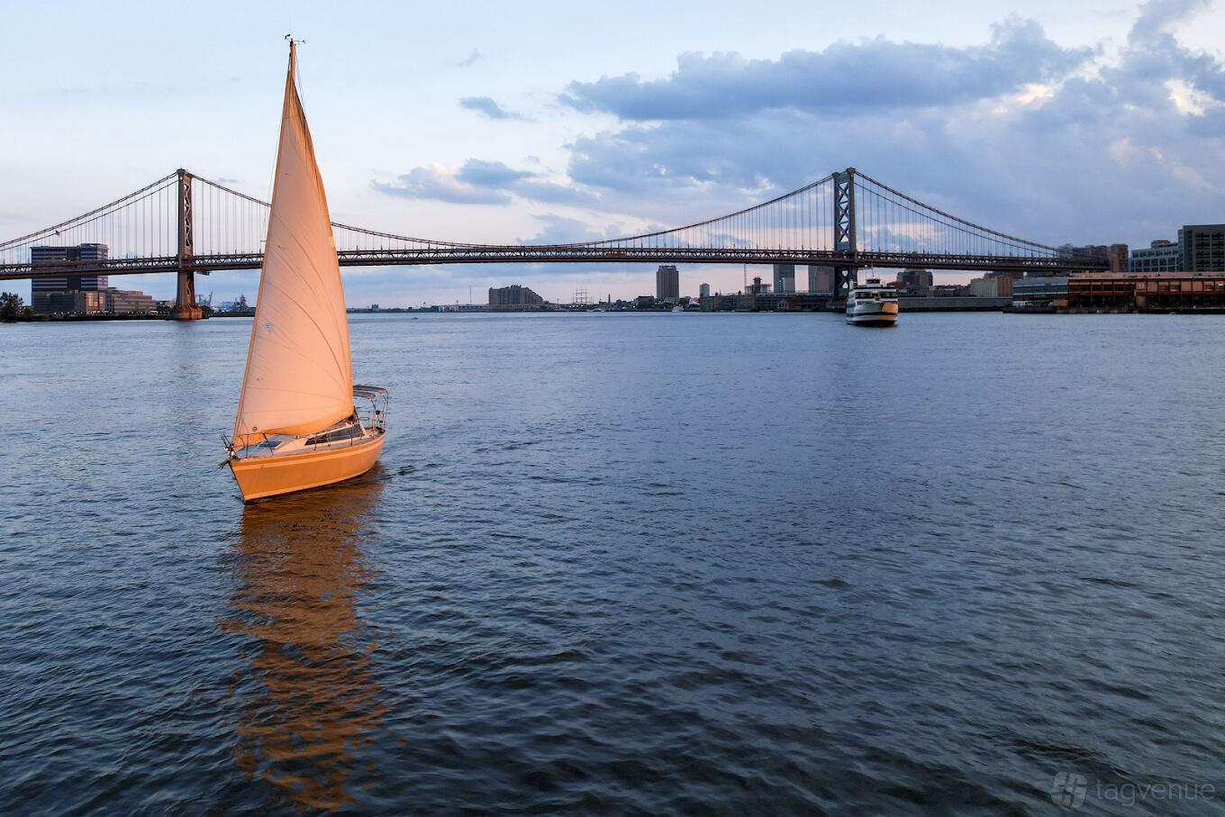 A sailboat with white sails on the river near a suspension bridge at Phila Sail.