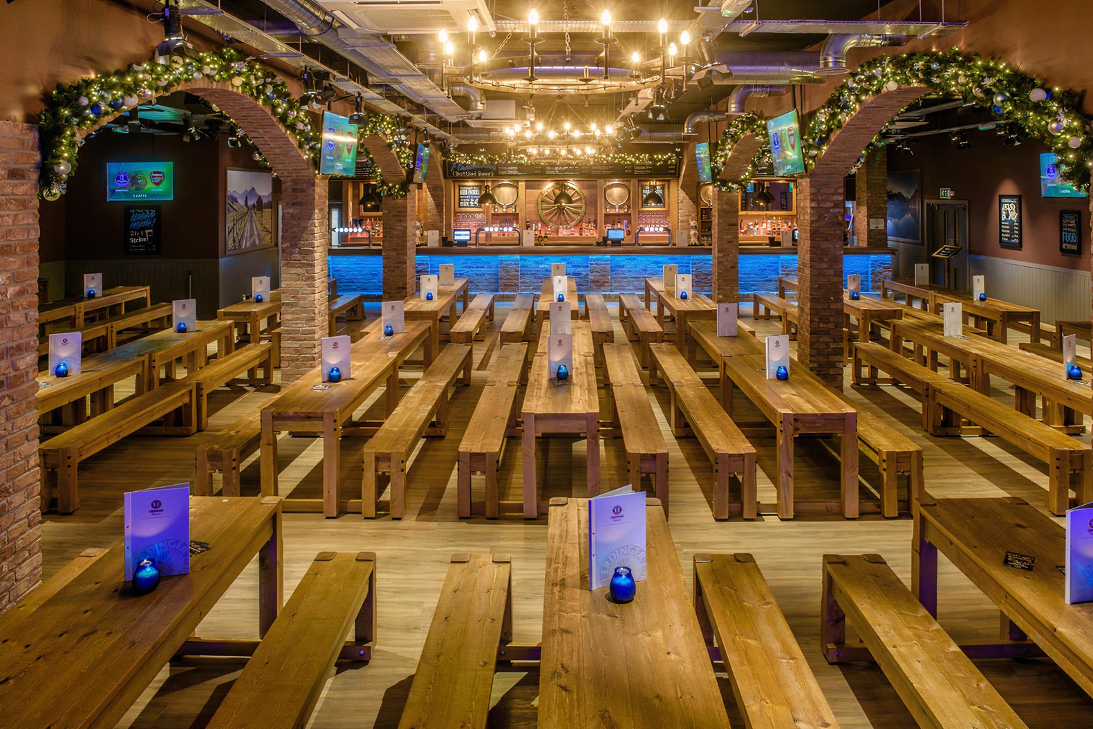 A bar with long wooden communal tables, exposed brick arches, and hanging Edison bulb lights at The Bierkeller Birmingham.
