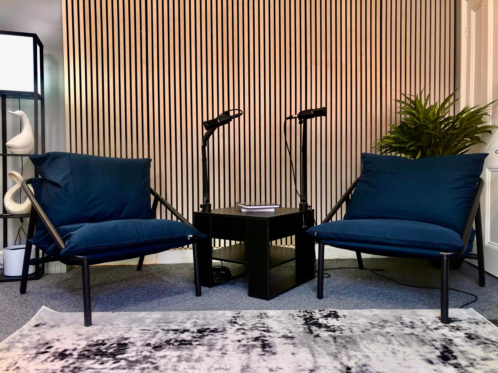 A recording room with acoustic slat wood paneling, two blue lounge chairs, microphones, and a small table at Leeds Content Creation Studios.