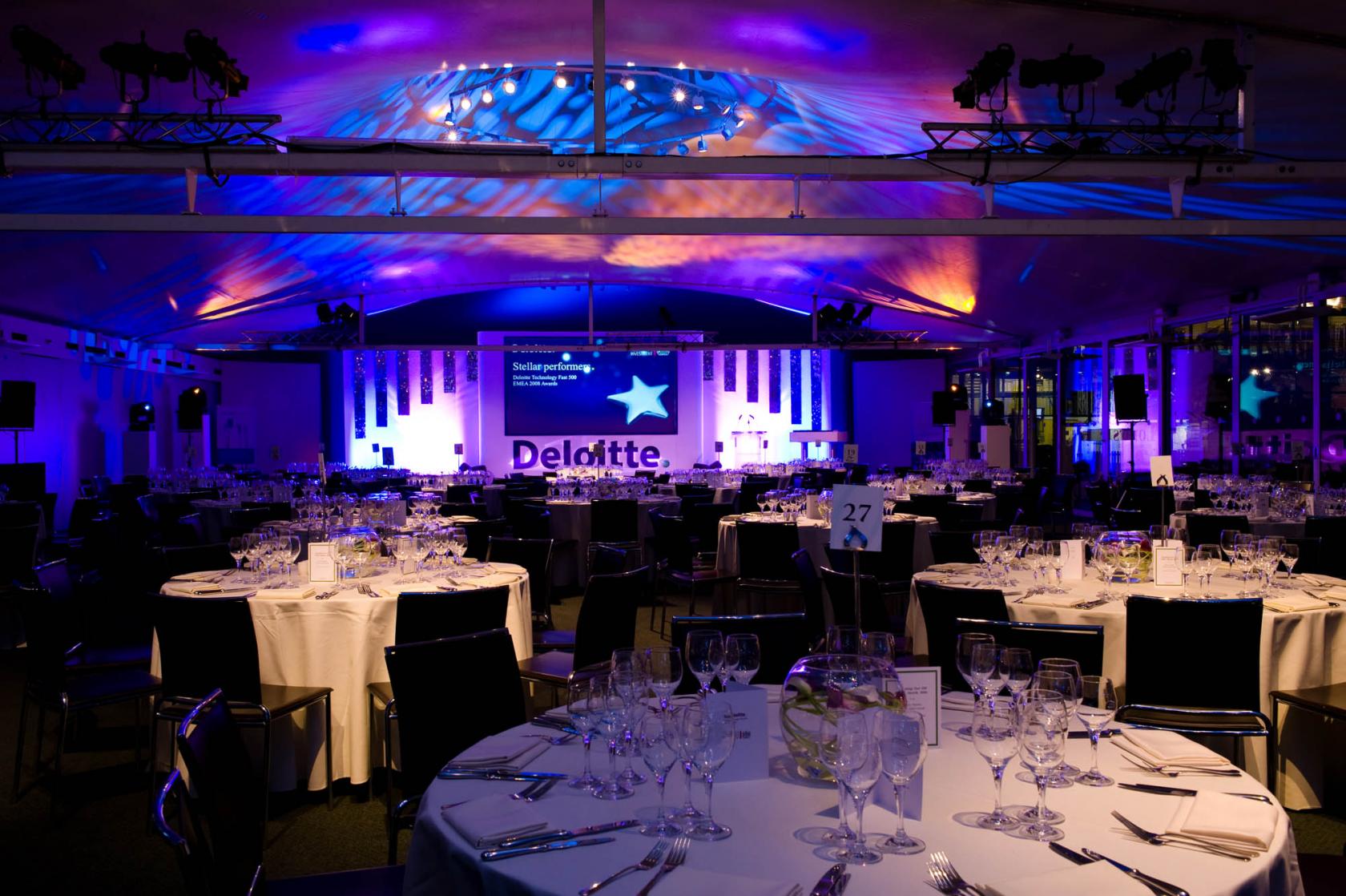 An event venue with round tables dressed in white linens, glassware, and purple accent lighting at Lord's Cricket Ground.
