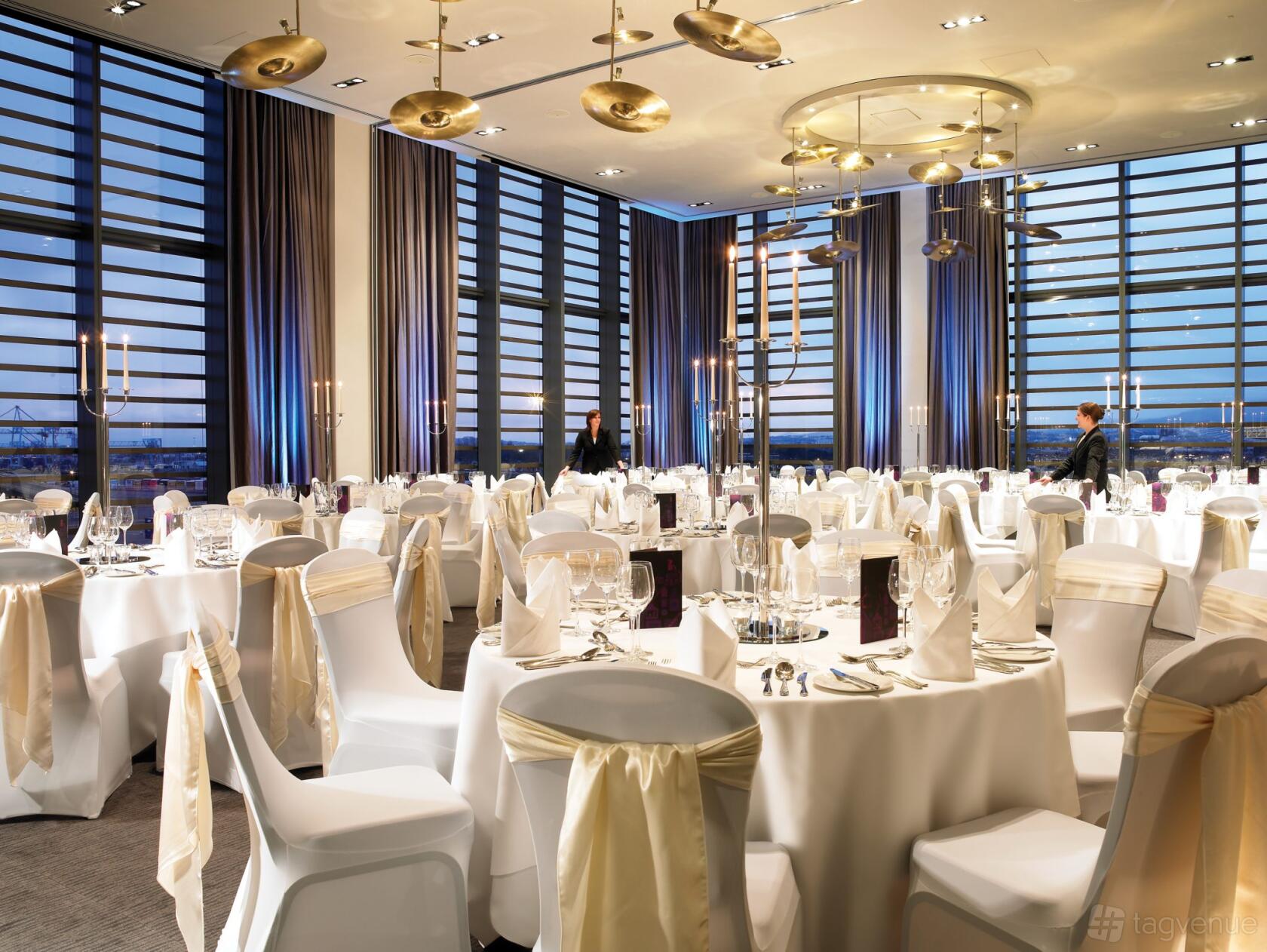 A hotel banquet hall with floor-to-ceiling windows, round tables with white linens, and gold chair sashes at The Gibson Hotel.