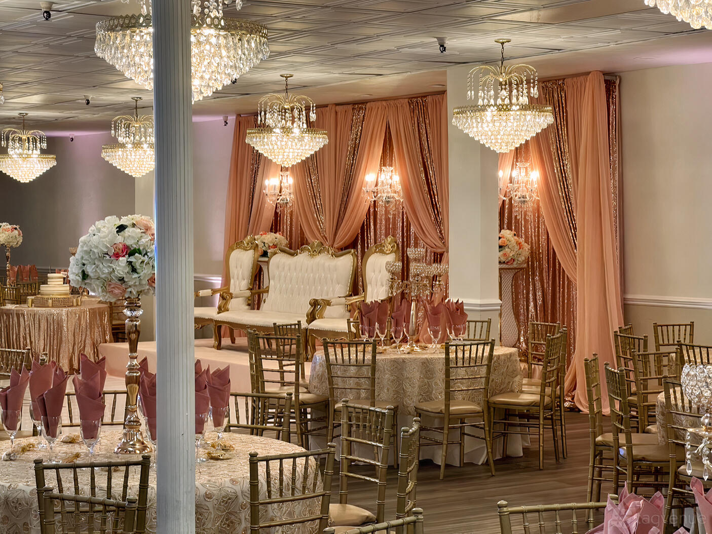 A banquet hall with gold chandeliers, peach drapery, and round tables dressed in lace linens at Pearl Banquet Hall.
