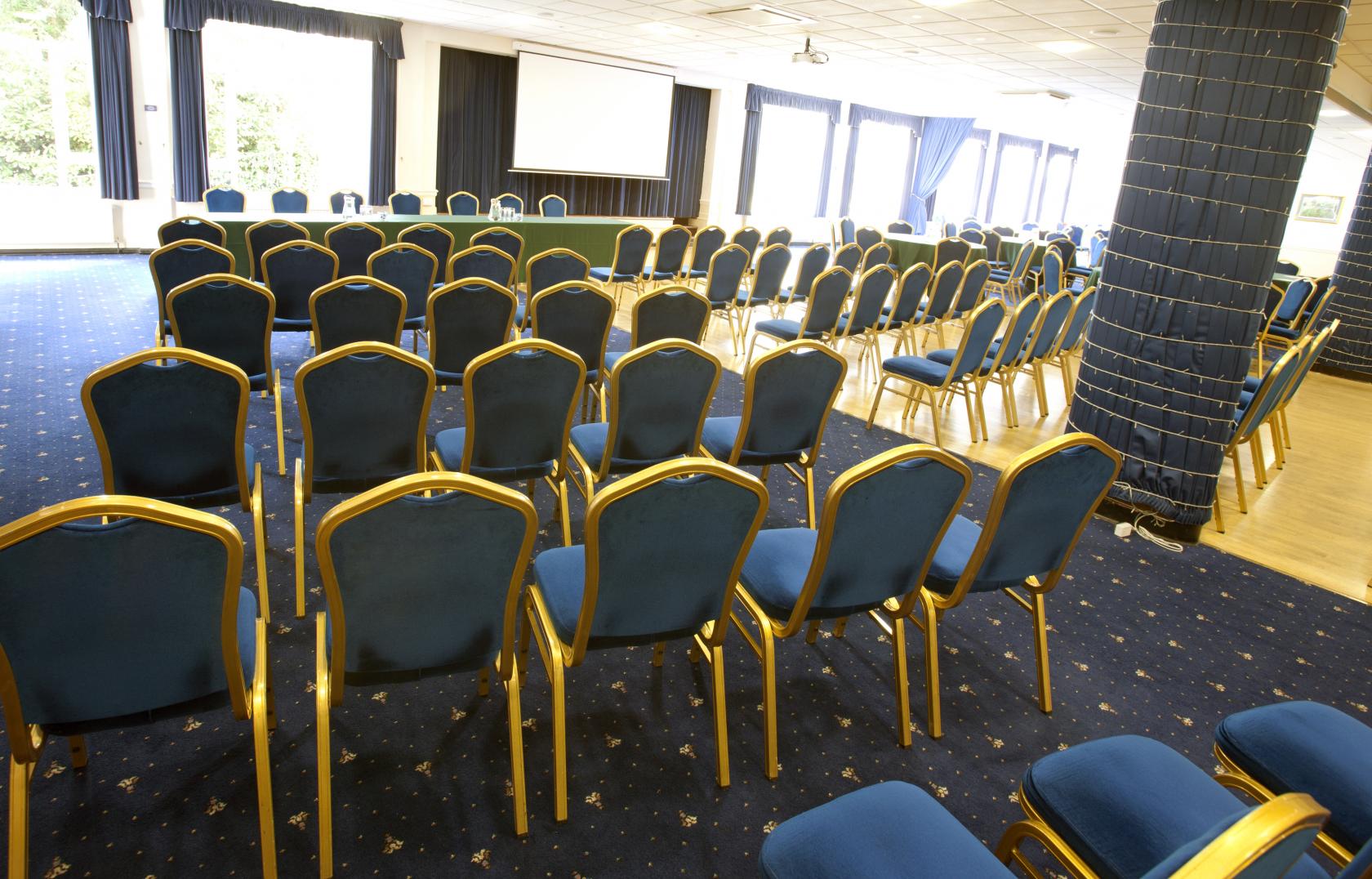 An event space with blue cushioned chairs, large windows, and a projector screen at Sindlesham Court.