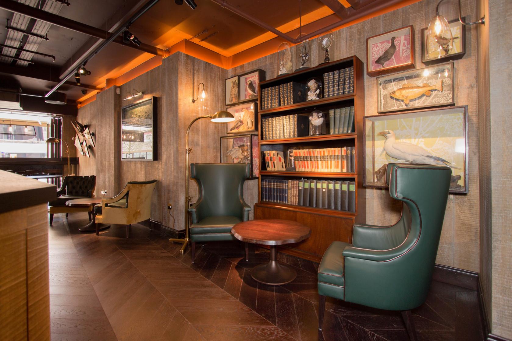A cocktail bar with wingback leather chairs, floor lamp, bookshelves, and wood-paneled walls at The Botanist Broadgate Circle.