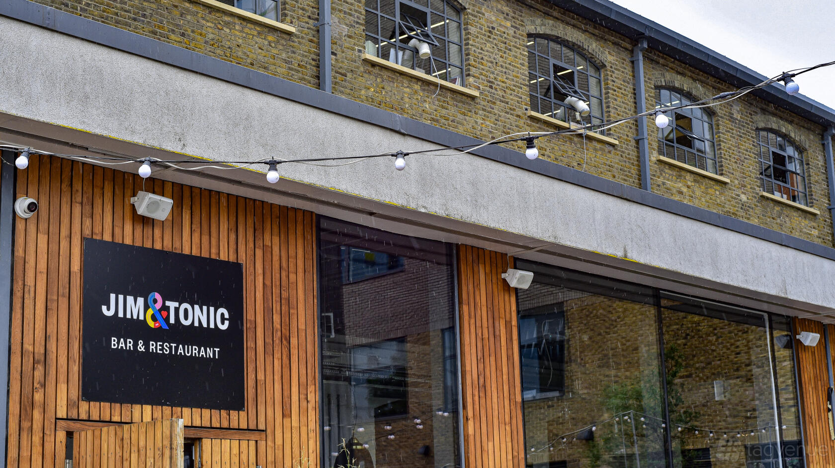 A pub exterior with exposed brick walls, large front windows, and string lights at The Printhouse at Jim & Tonic East.
