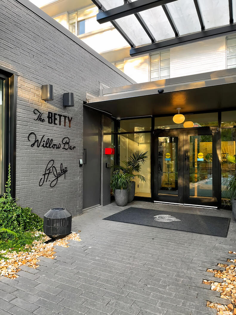 A restaurant entrance with glass double doors, potted plants, and a gray brick wall at The Betty.