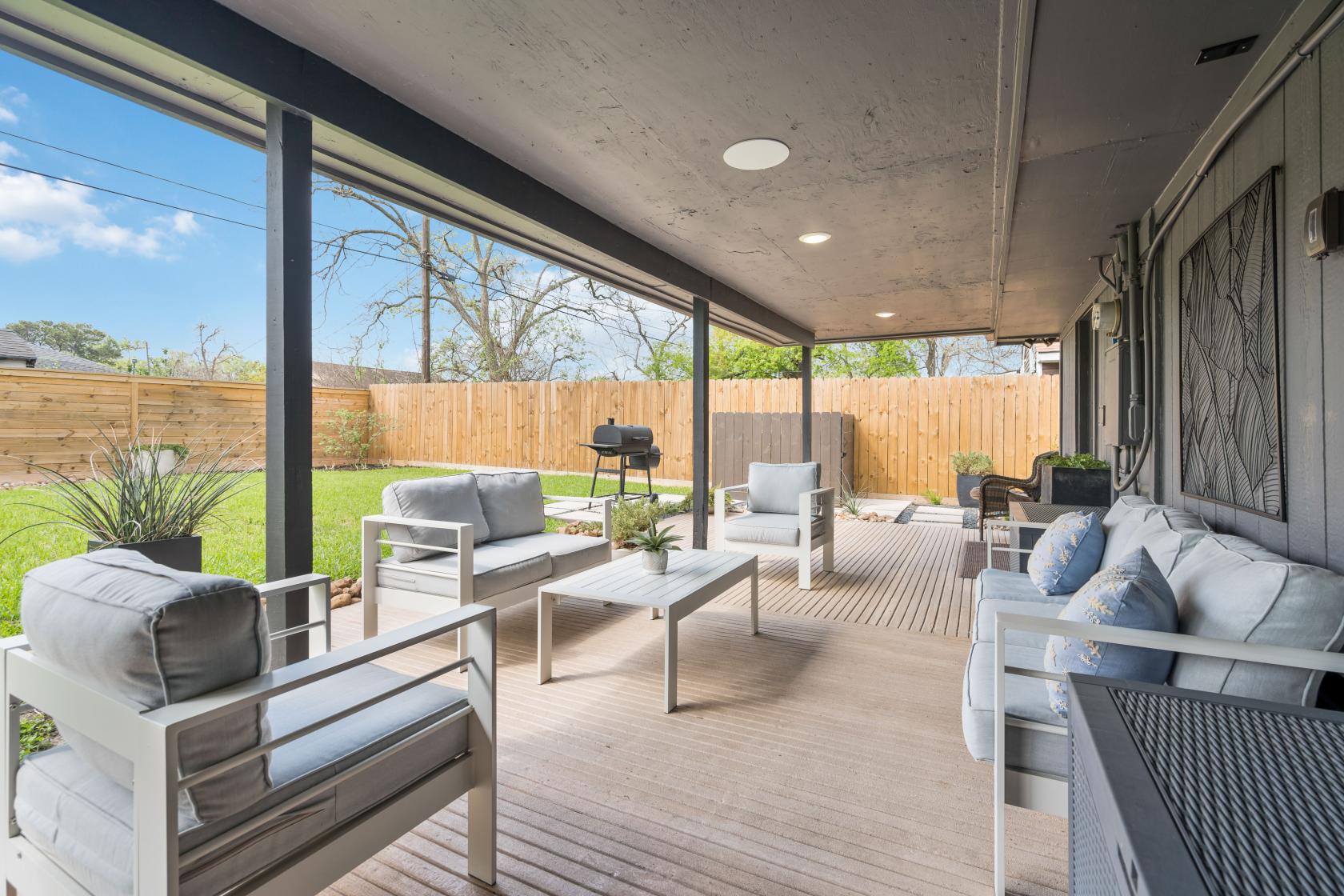 A covered patio with cushioned lounge chairs, potted plants, and a grill overlooking a private yard at The Modern Haven Home.