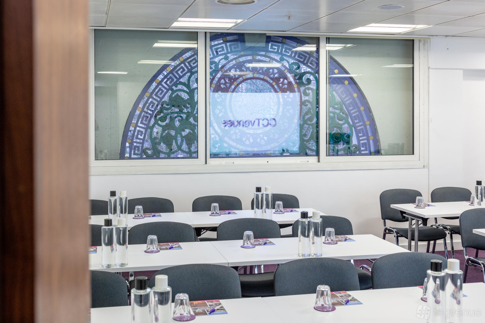 A conference centre with rows of tables, grey chairs, bottled water, and a large decorative window at CCT Venues - Smithfield.