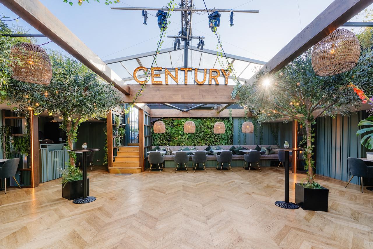 A terrace bar with a glass roof, herringbone wood floors, potted trees, and rattan pendant lighting at Century Club.