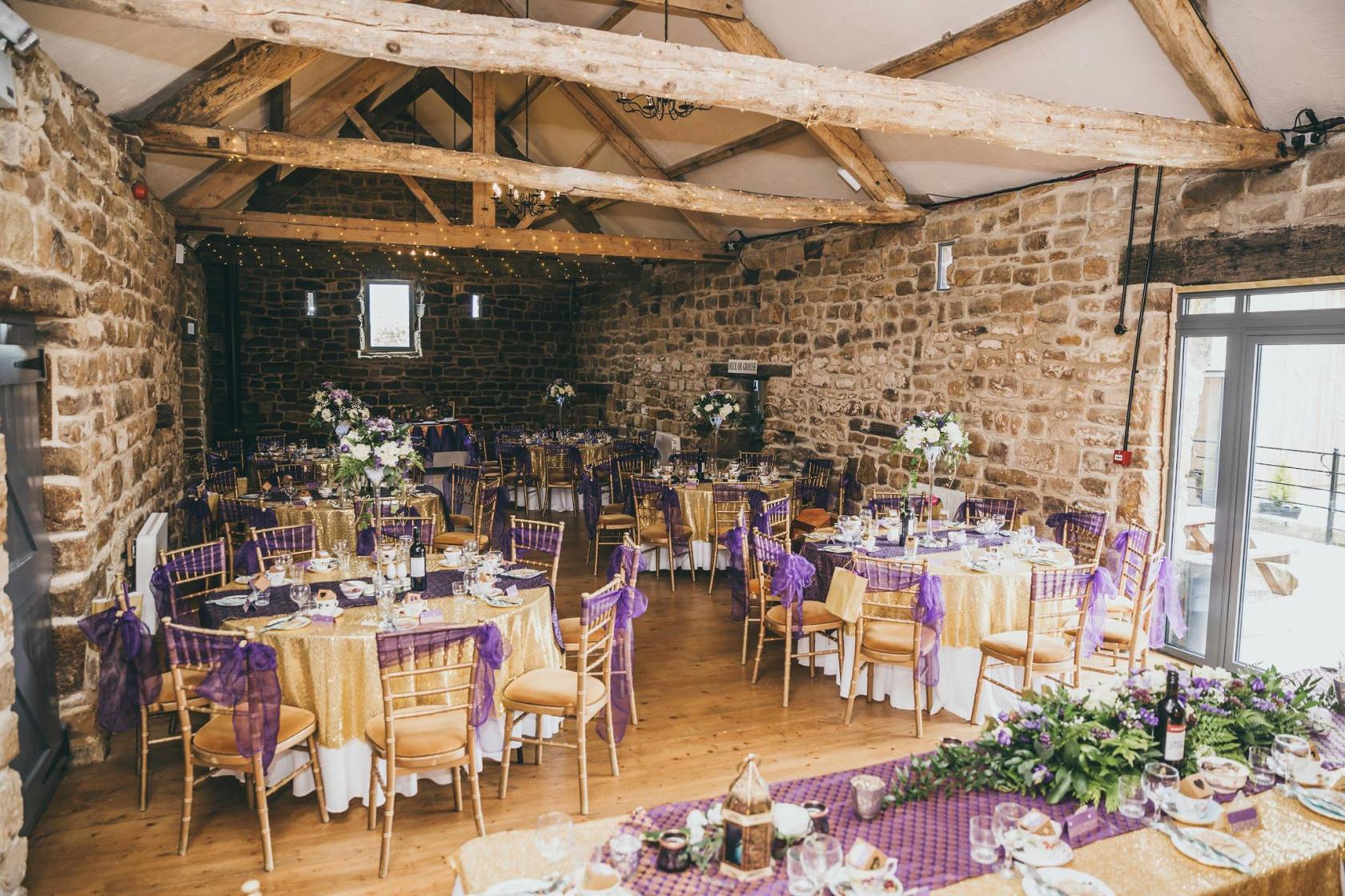 A castle event hall with exposed stone walls, wooden beams, and round tables with purple and gold linens at Danby Castle.