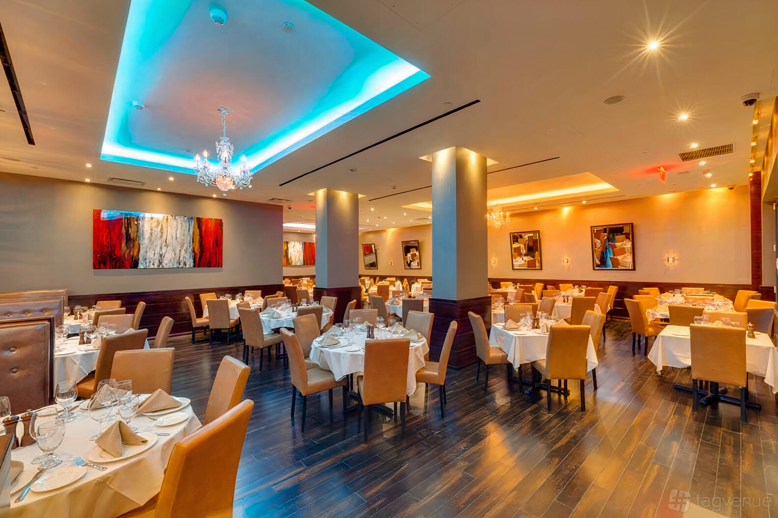 A restaurant dining area with tan upholstered chairs, round tables with white linens, chandeliers, and modern art at Empire Steak House West.