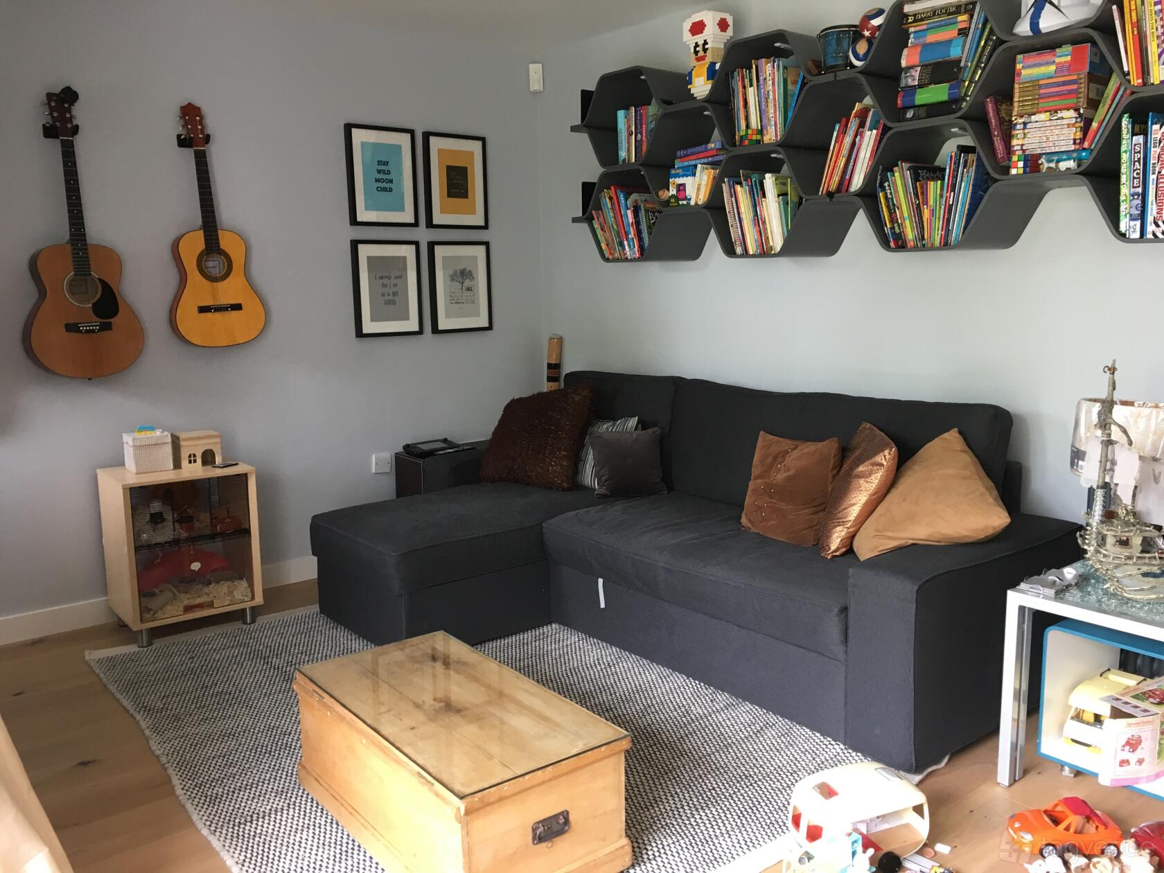 A living room with a gray sectional sofa, wall-mounted bookshelves, guitars, and framed art at Whitelands Crescent.