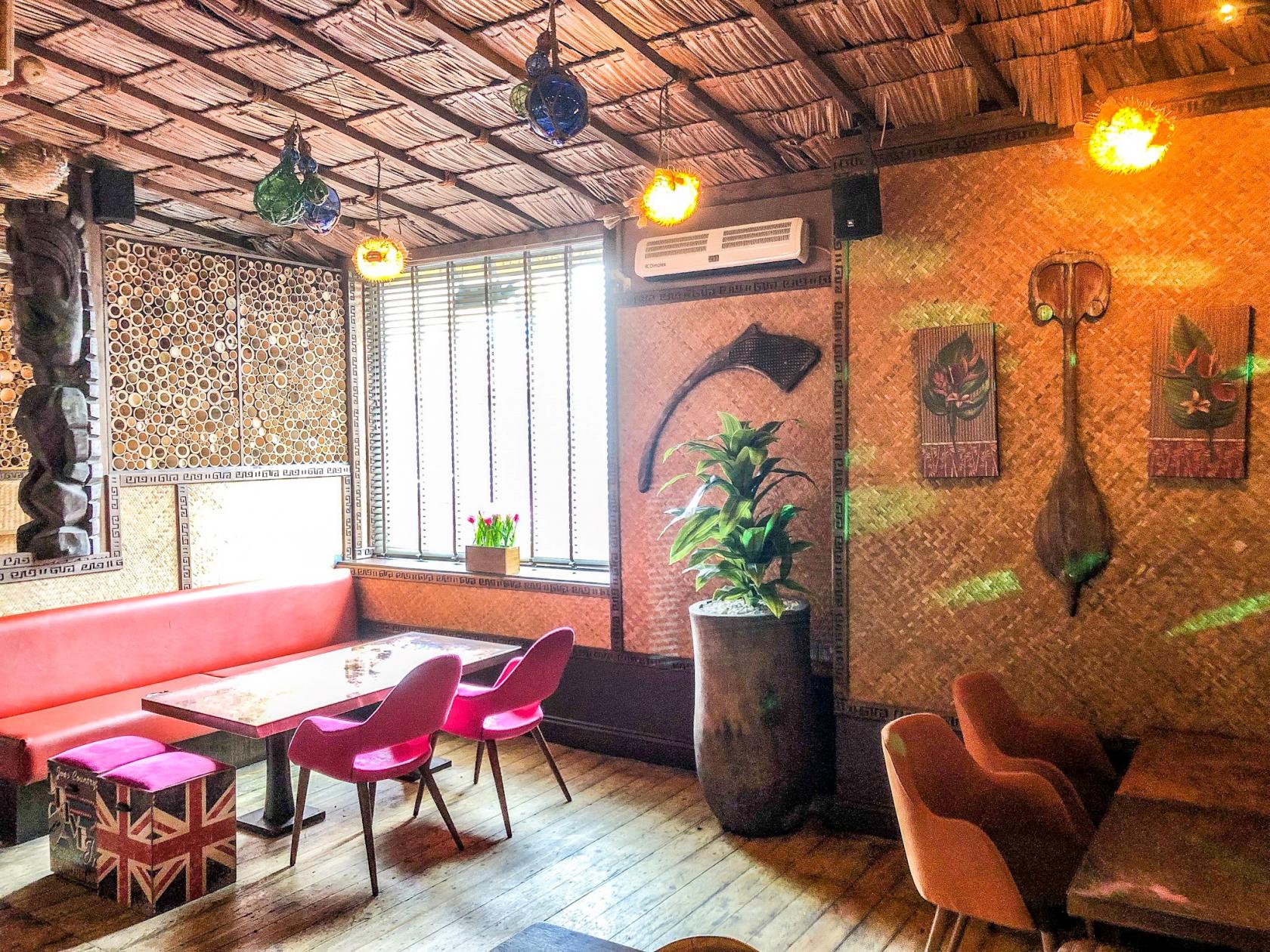 A lounge in a cocktail bar with bamboo walls, pink chairs, a red cushioned bench, and tropical decor at Kona Kai.