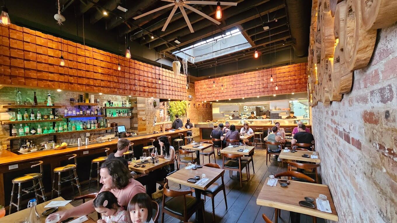A restaurant with wooden tables, exposed brick walls, and a skylight ceiling at Bushniwa.