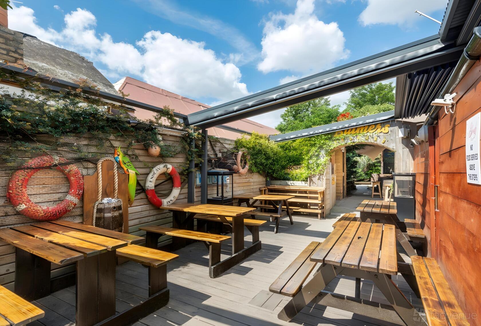 An outdoor pub area with wooden picnic tables, wall-mounted lifebuoys, and overhead pergola at The Bloody Stream.