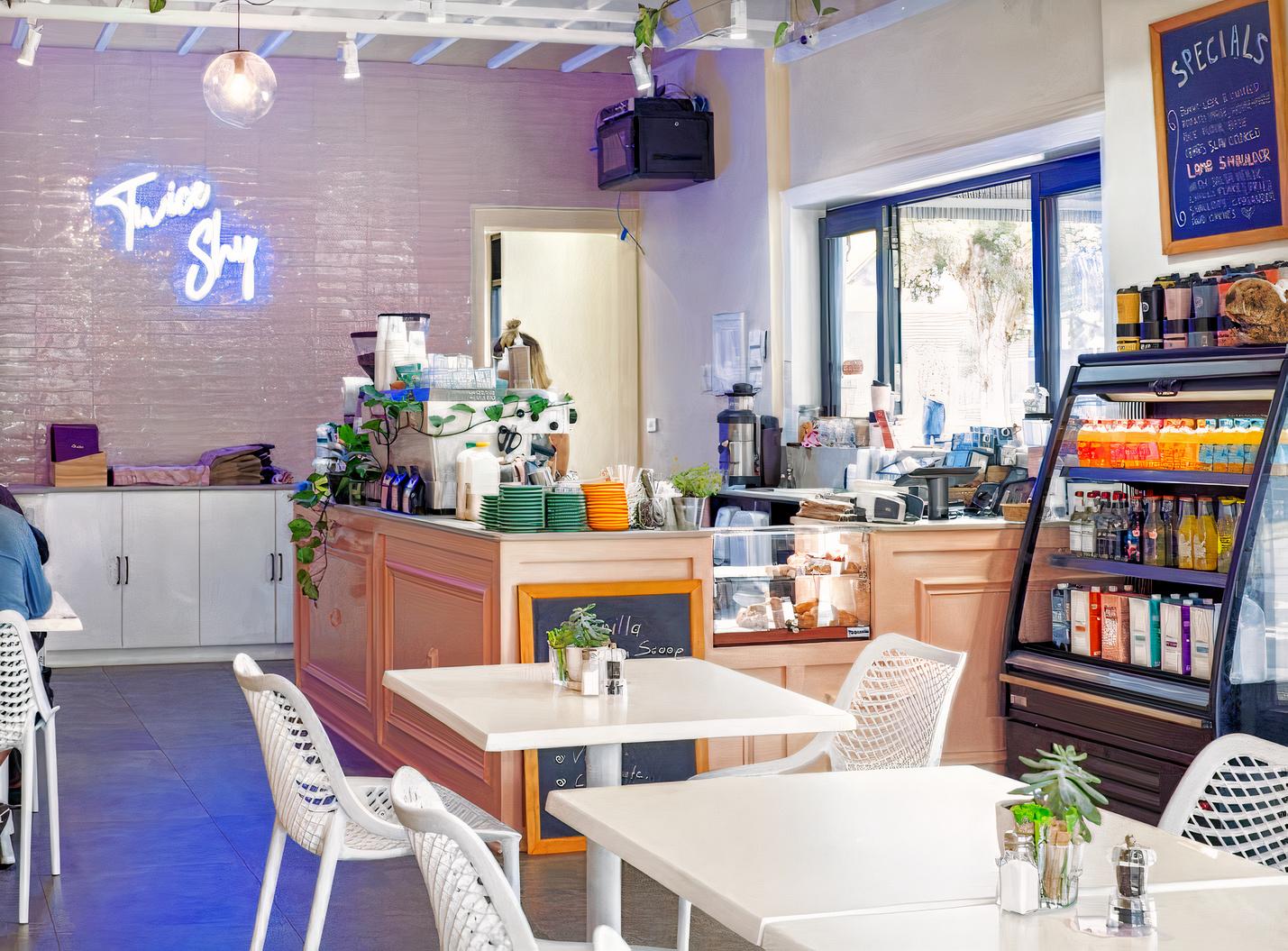 A cafe with white tables, a pastel counter, neon signage, and a glass display fridge at Twice Shy.