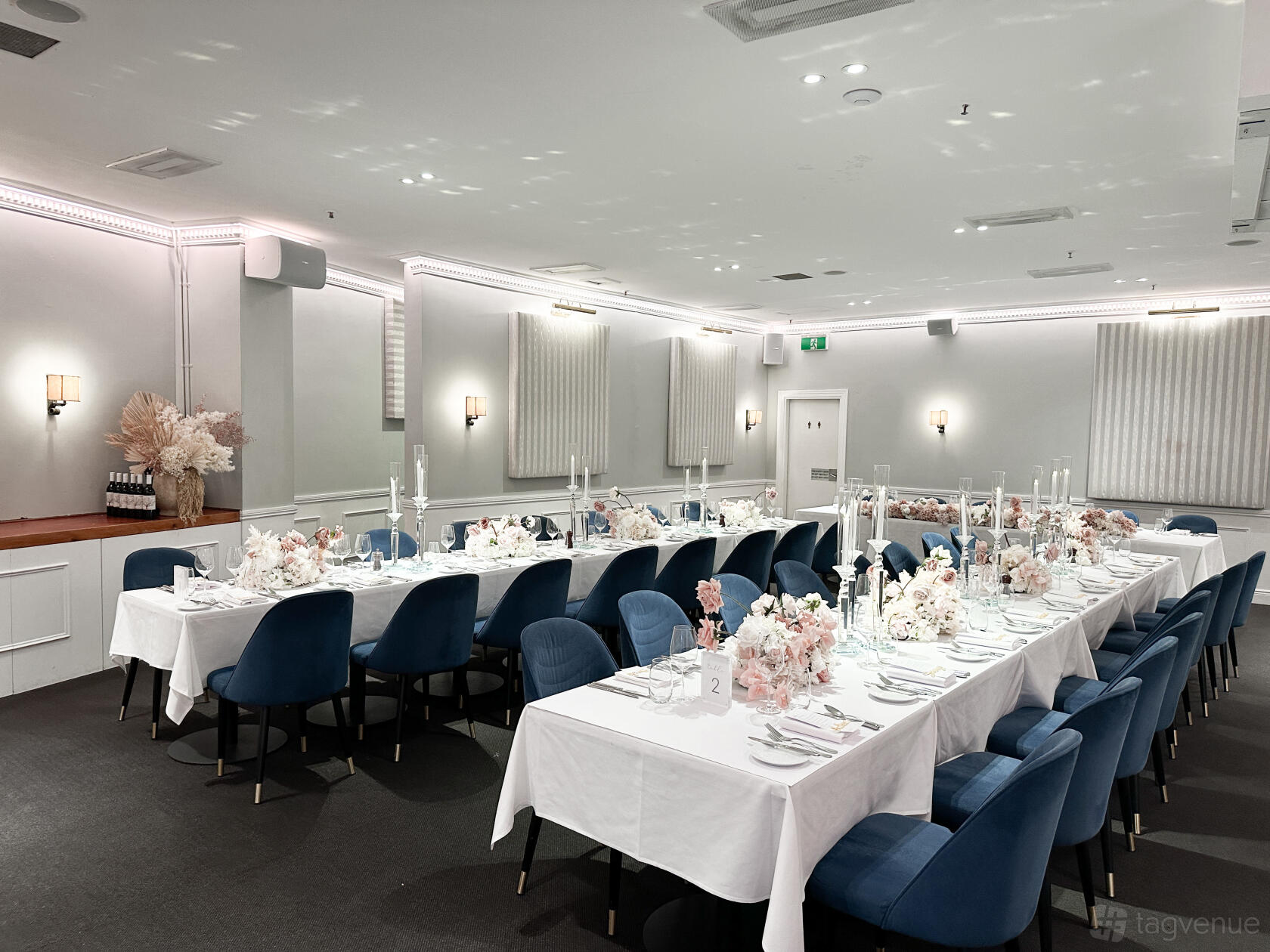A dining room with long tables covered in white linens, blue velvet chairs, and floral centerpieces at The Royal Exchange of Sydney.