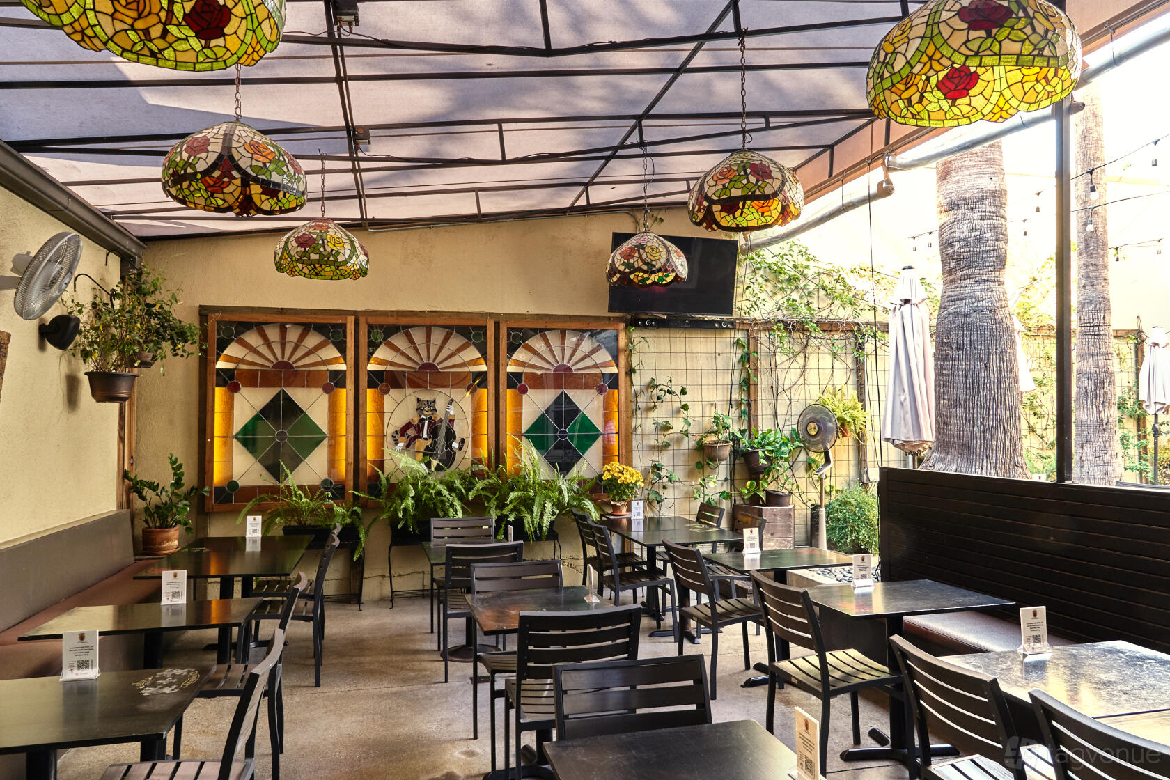 A terrace with stained glass panels, hanging Tiffany-style lamps, and black tables at The Cat & Fiddle Pub & Restaurant.