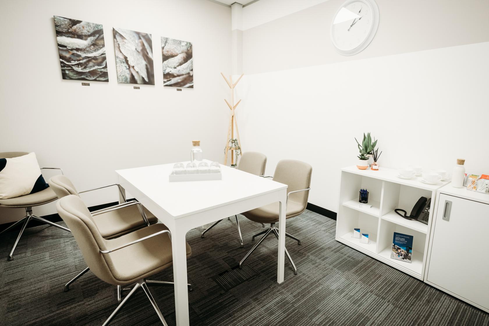 A meeting room with a white table, four beige chairs, wall art, and a small shelving unit at BusinessLodge Stoke.