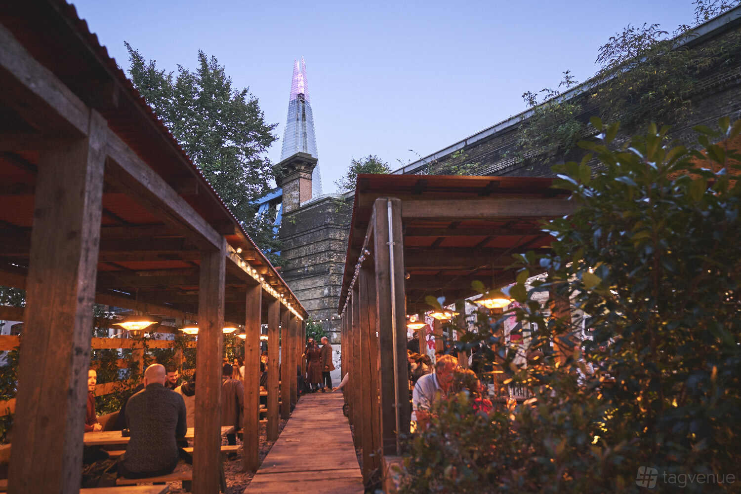 An outdoor railway arch bar with wooden pergolas, string lights, and The Shard visible at Flat Iron Square.