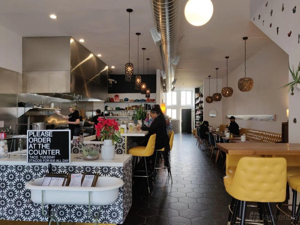A restaurant with an open kitchen, hexagon tile floor, yellow chairs, and pendant lights at Tato.