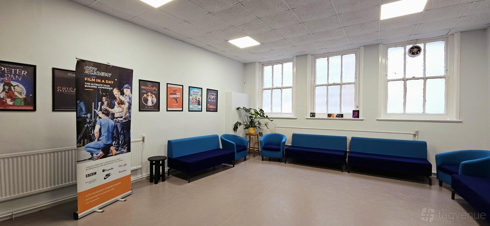 A rehearsal room with blue sofas, large windows, poster displays, and a potted plant at City Academy at 10 Herbal Hill.