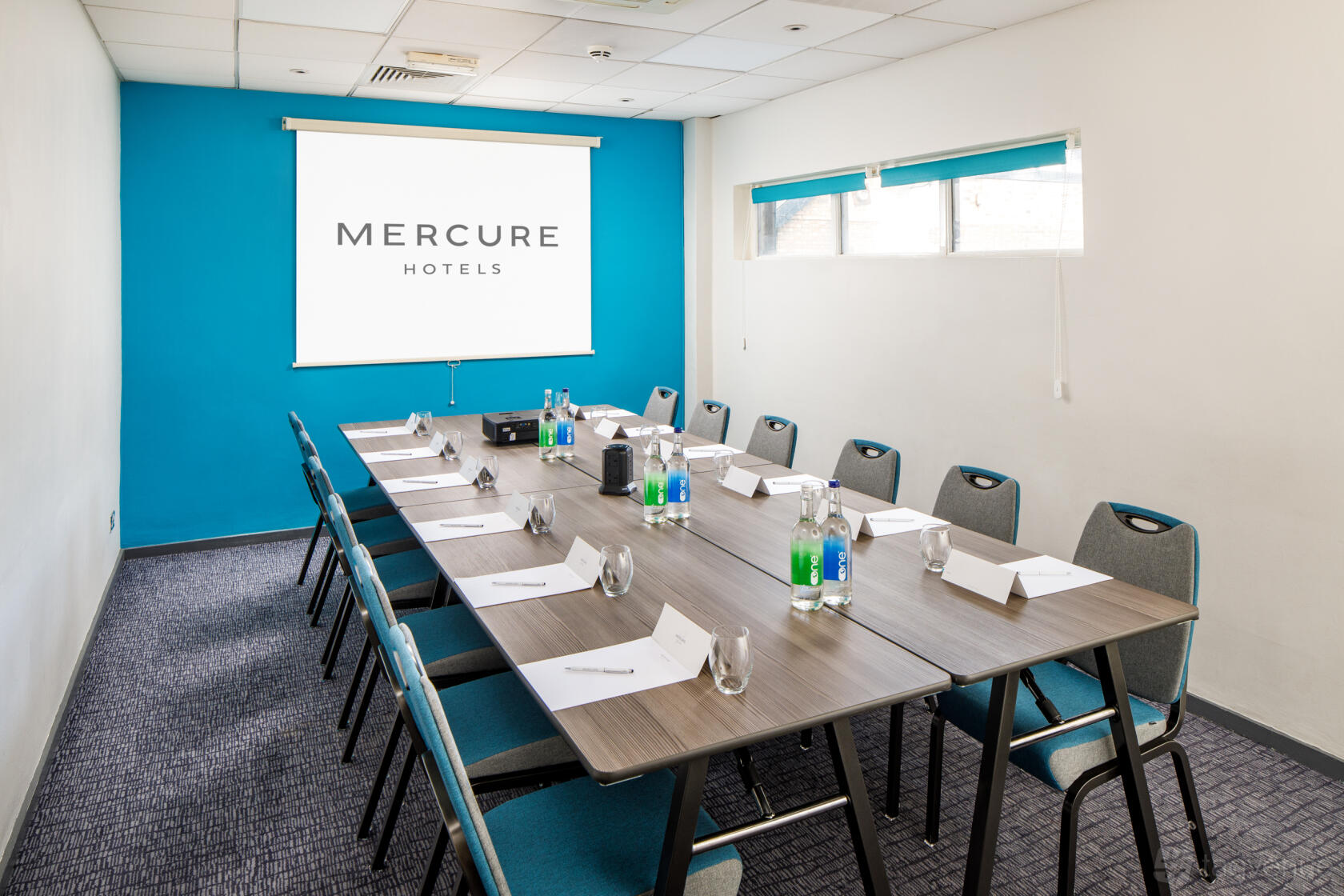A meeting room with a long conference table, projection screen, and bottled water at Mercure Bedford Centre.