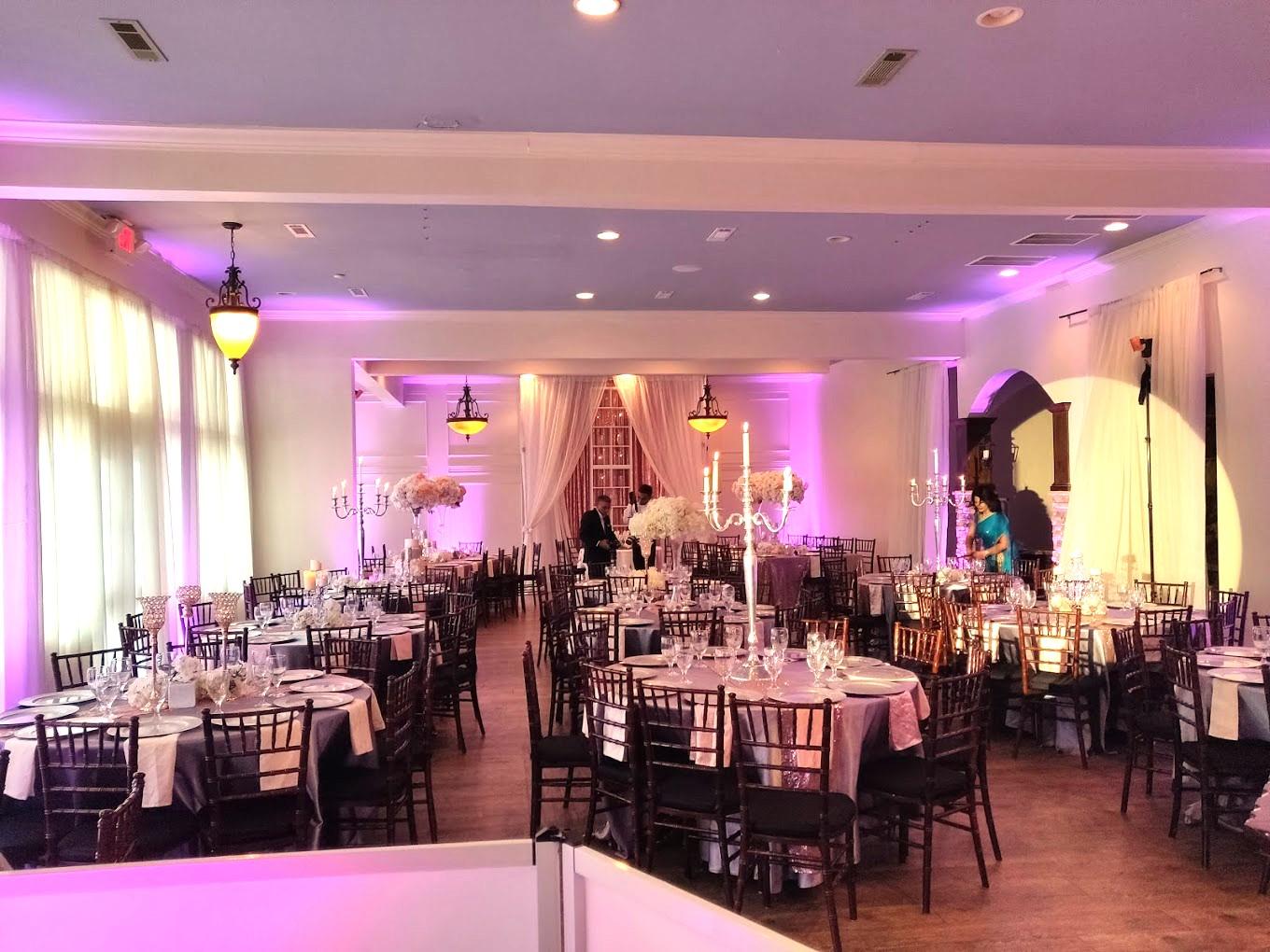 An event venue with round tables draped in white linens, tall floral centerpieces, and purple uplighting at Orleangham Event Facility.