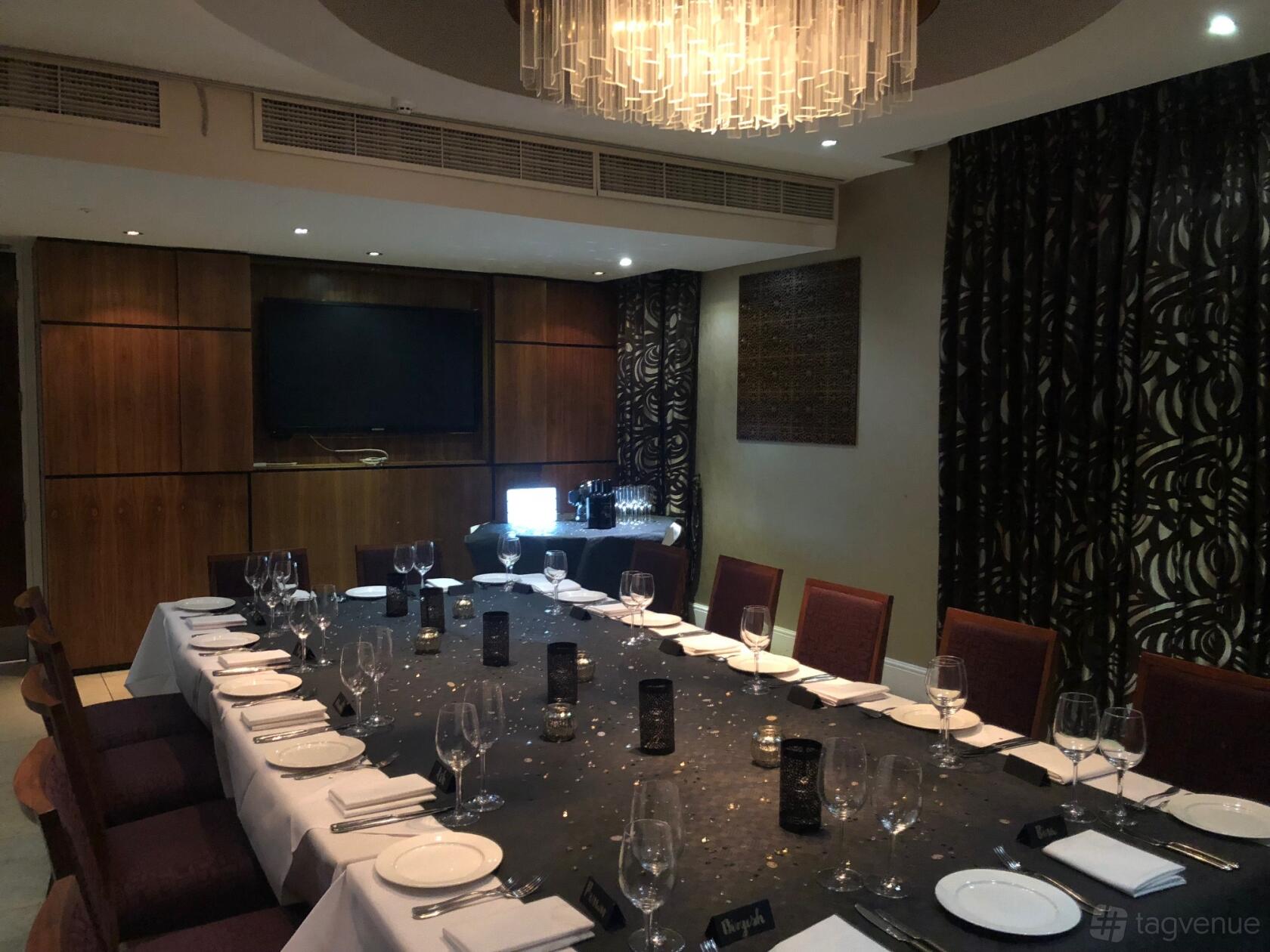 A dining room with a long banquet table, dark patterned curtains, and a wall-mounted TV at Bombay Palace.