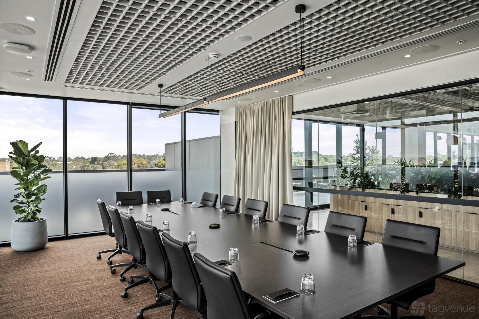 A boardroom with a long conference table, black chairs, floor-to-ceiling windows, and indoor plants at Hub Local Box Hill.