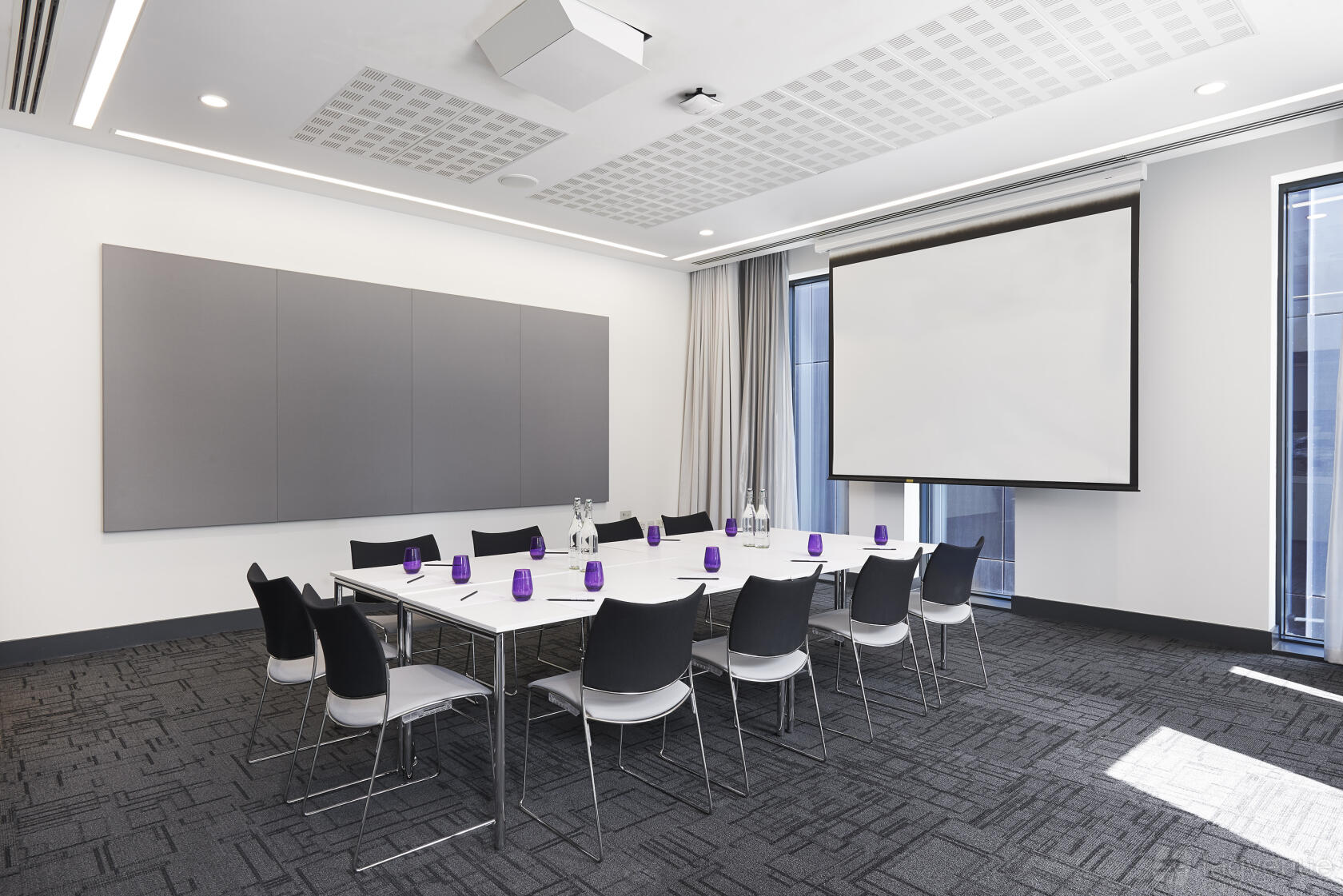 A meeting room with a square table, black chairs, water glasses, and a projection screen at INNSIDE Manchester.