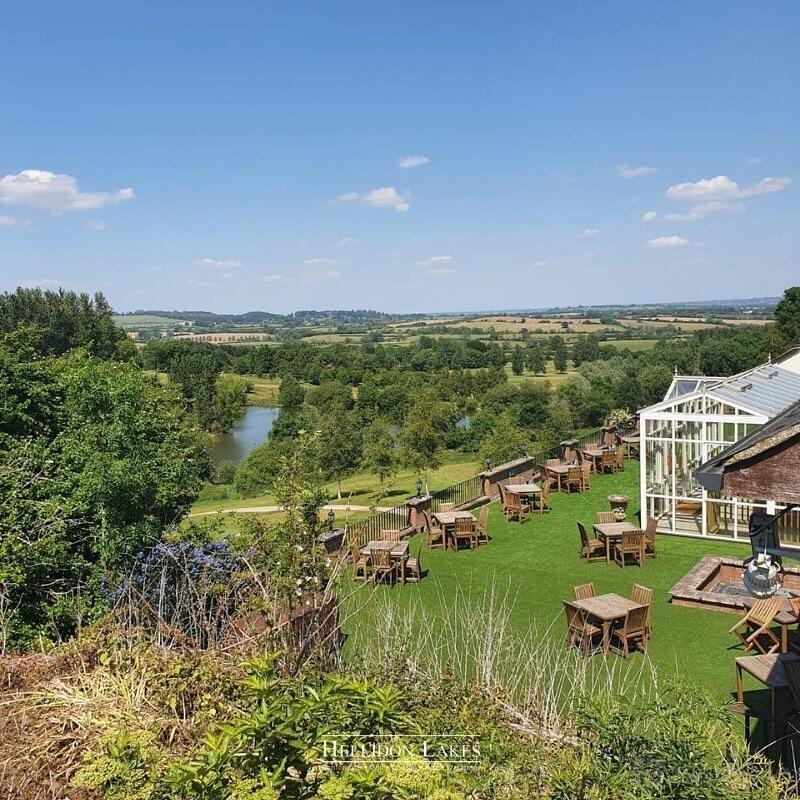 A hotel with outdoor wooden tables on a lawn overlooking scenic countryside and a lake at Hellidon Lakes Hotel.