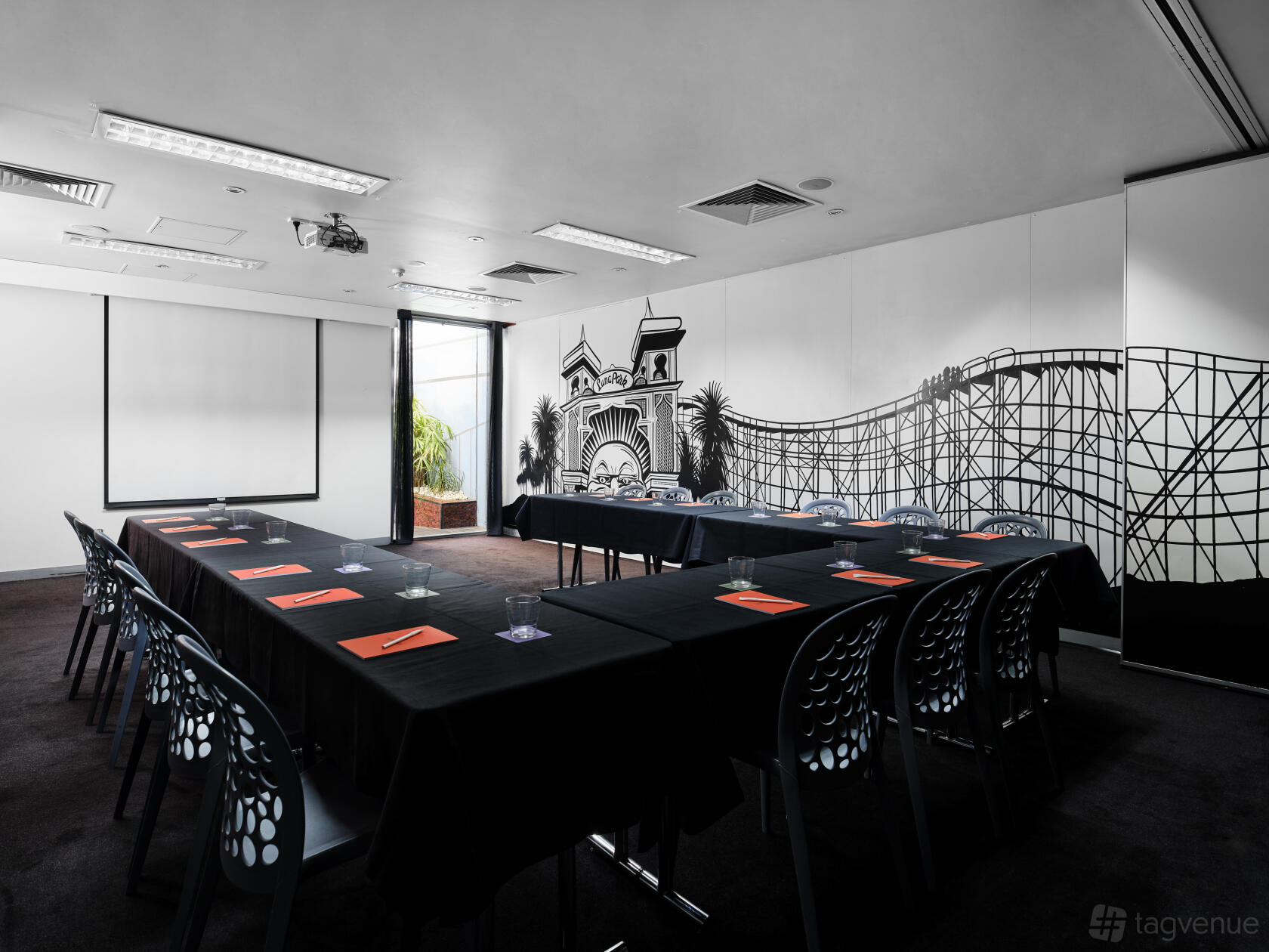 A meeting room with a mural of Luna Park, natural light, black tablecloths, and U-shaped seating at Saint Kilda Beach Hotel.