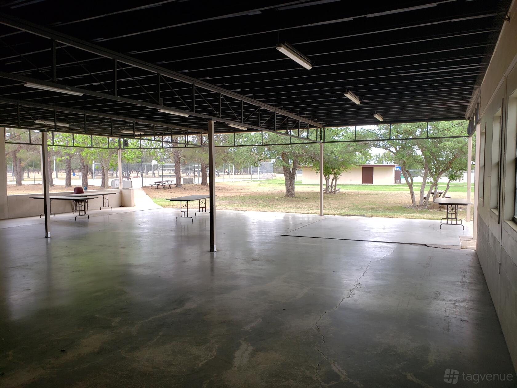 An outdoor covered patio with concrete flooring, open sides, and scattered folding tables at Texas 4-H Conference Center.