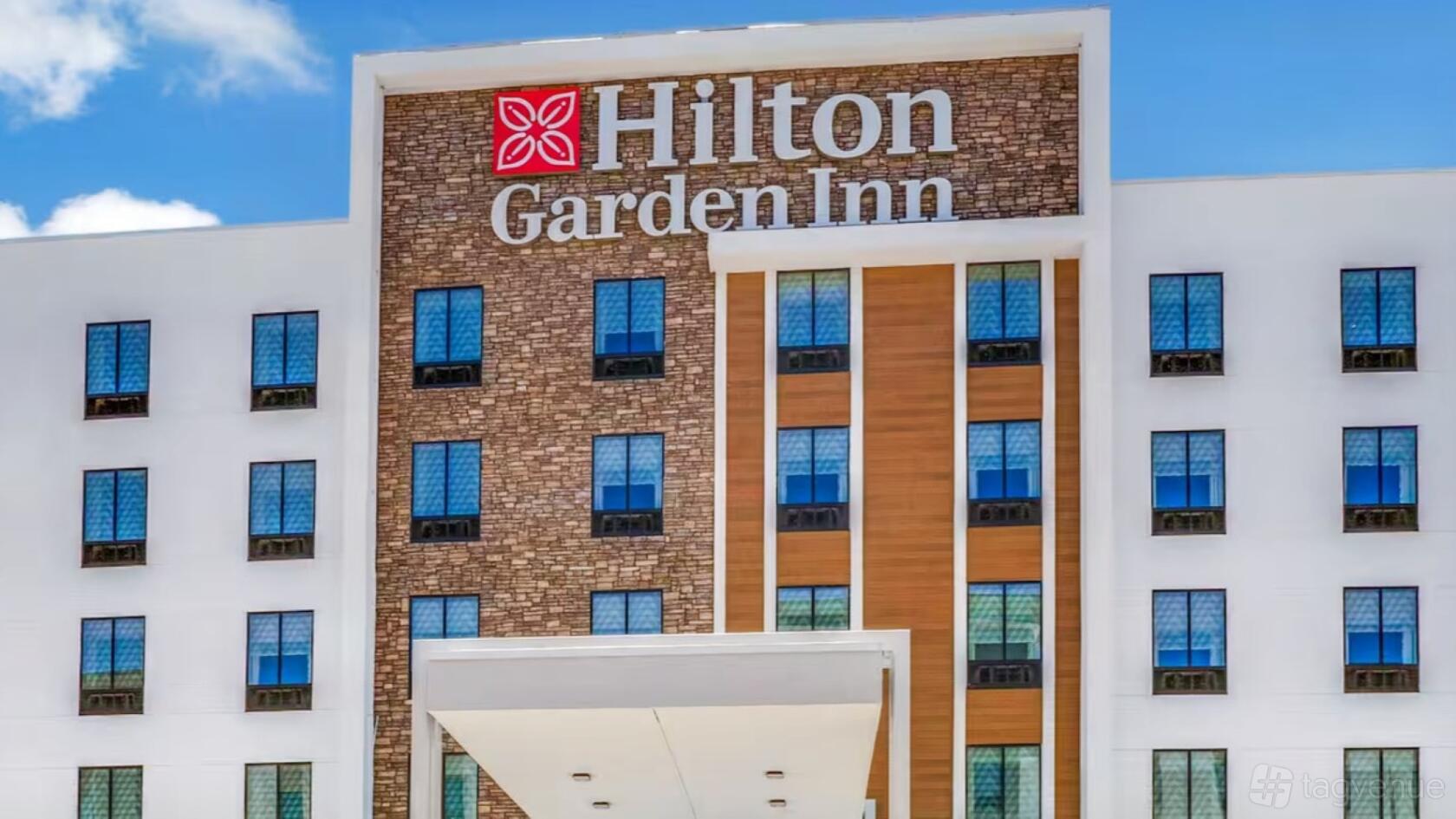 A hotel exterior with a large Hilton Garden Inn sign, brick facade, and rows of guest room windows.