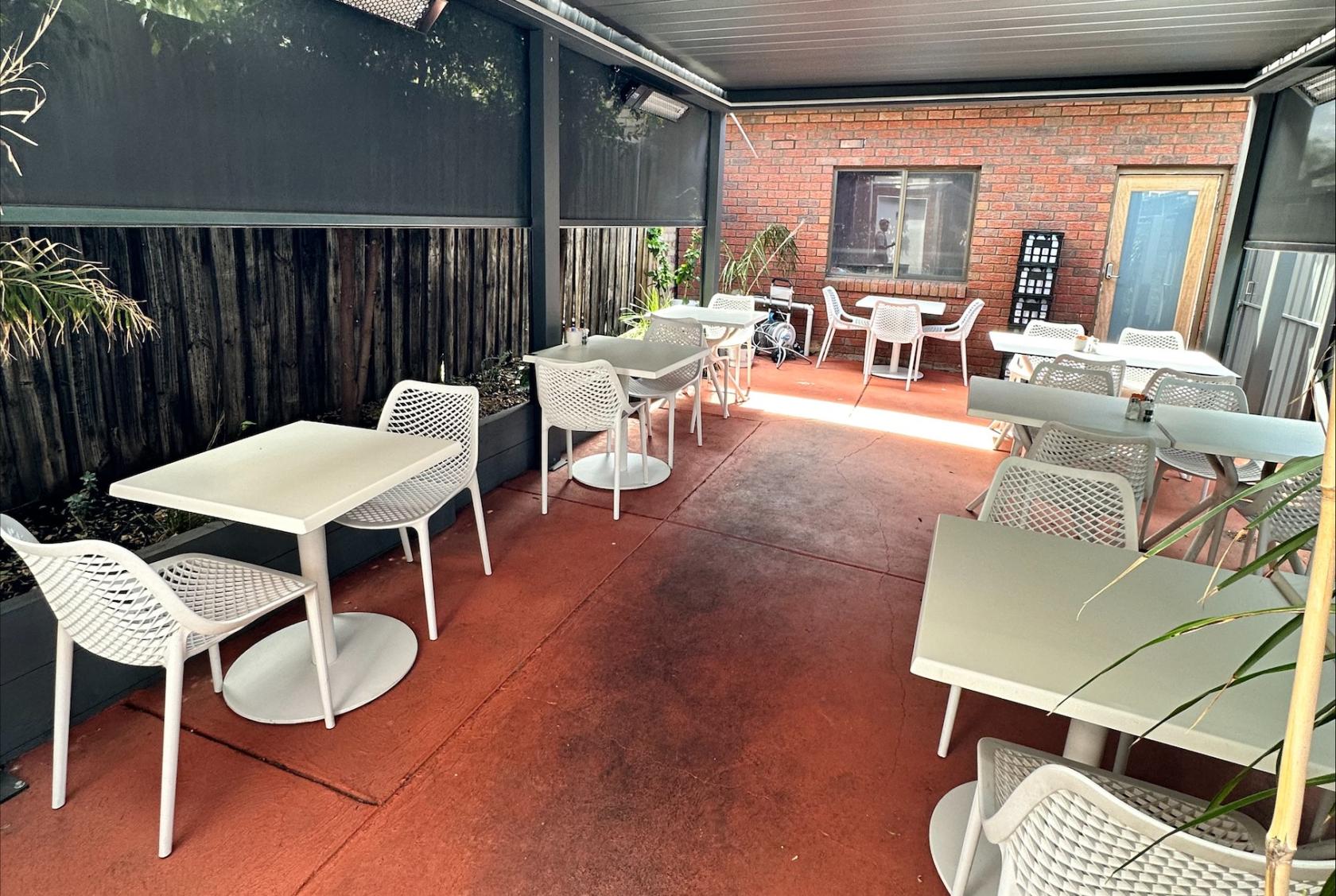 An outdoor cafe area with white mesh chairs, square tables, and partial covering at Twice Shy.