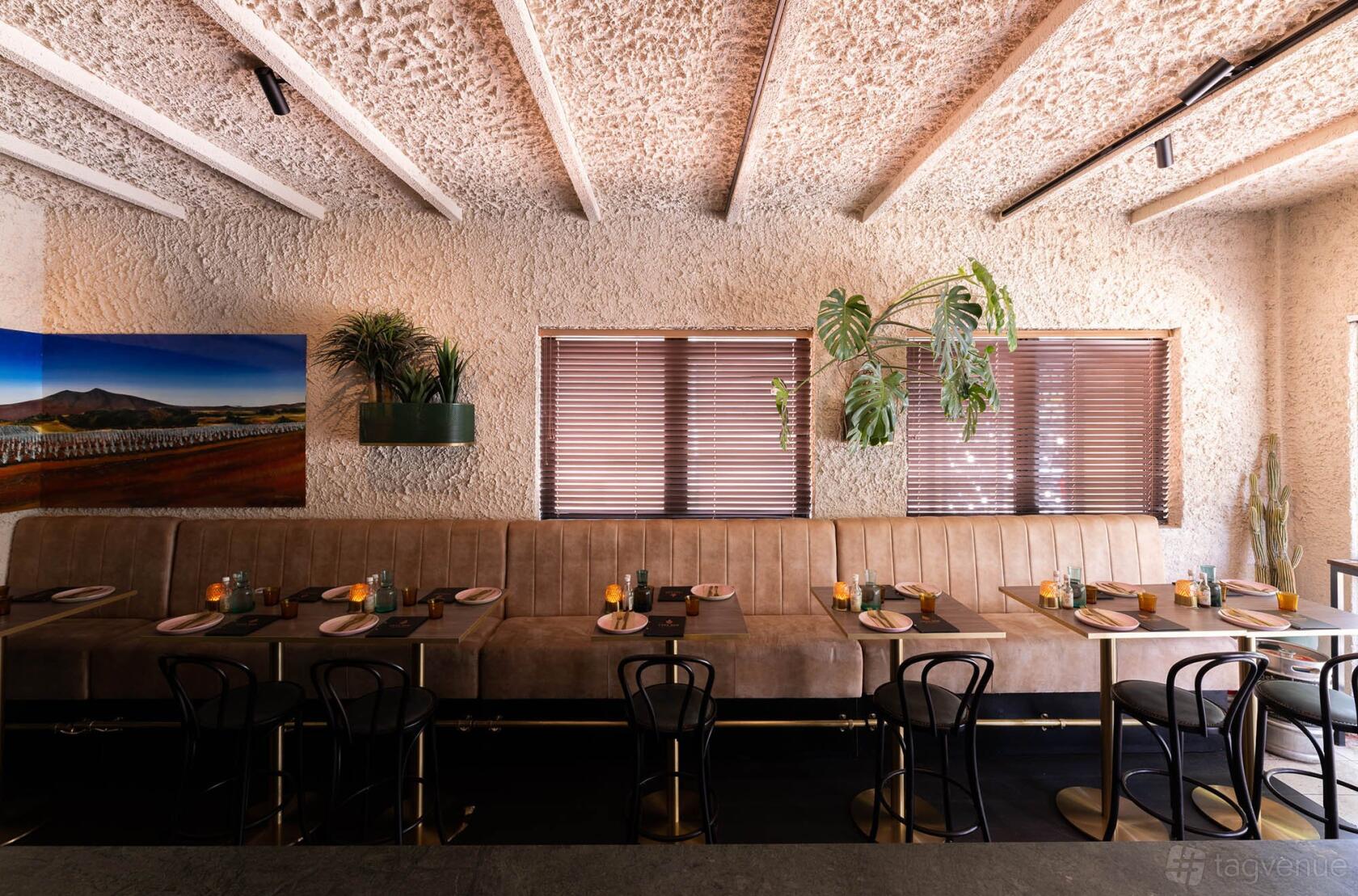 A restaurant with textured walls, wooden ceiling beams, tan banquette seating, and set tables at Casa N.O.M Bar y Restaurante.