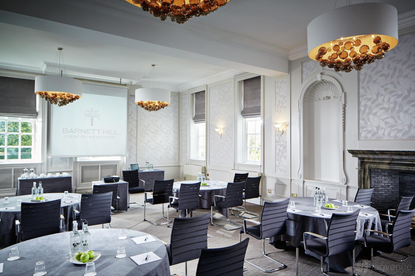 A function room with round tables, bottled water, modern chandeliers, and large windows at Barnett Hill Hotel.