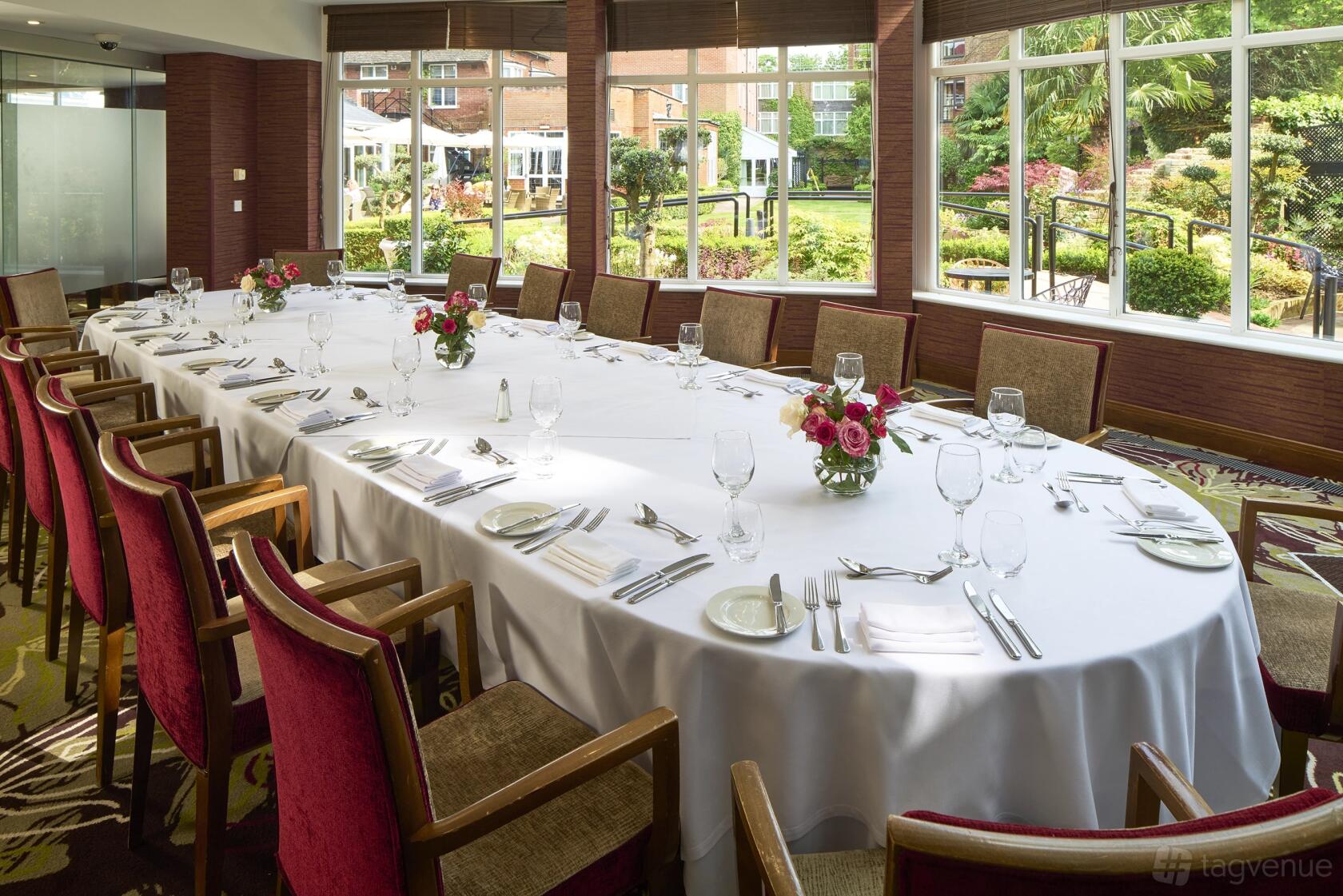 A hotel dining room with large windows overlooking gardens, set with a long oval table and red chairs at The Bull Hotel.