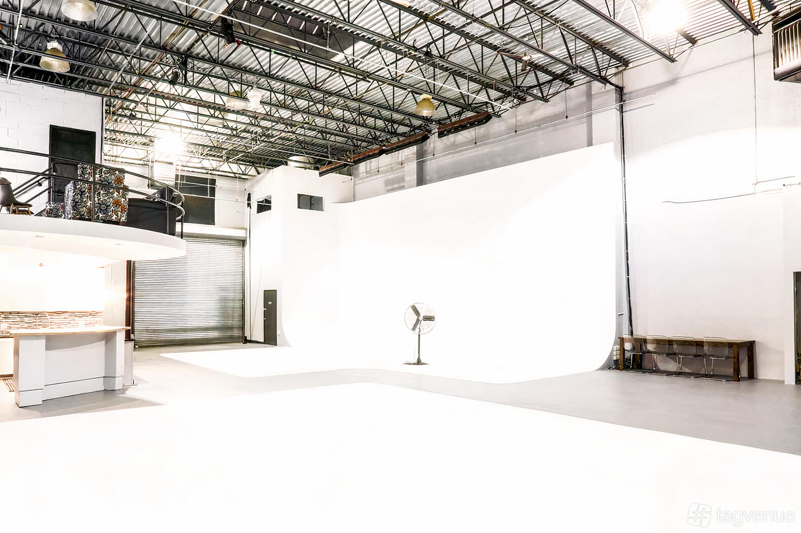 A spacious photo studio with a large white cyclorama wall, exposed ceiling beams, and a standing fan at 718 Studios.