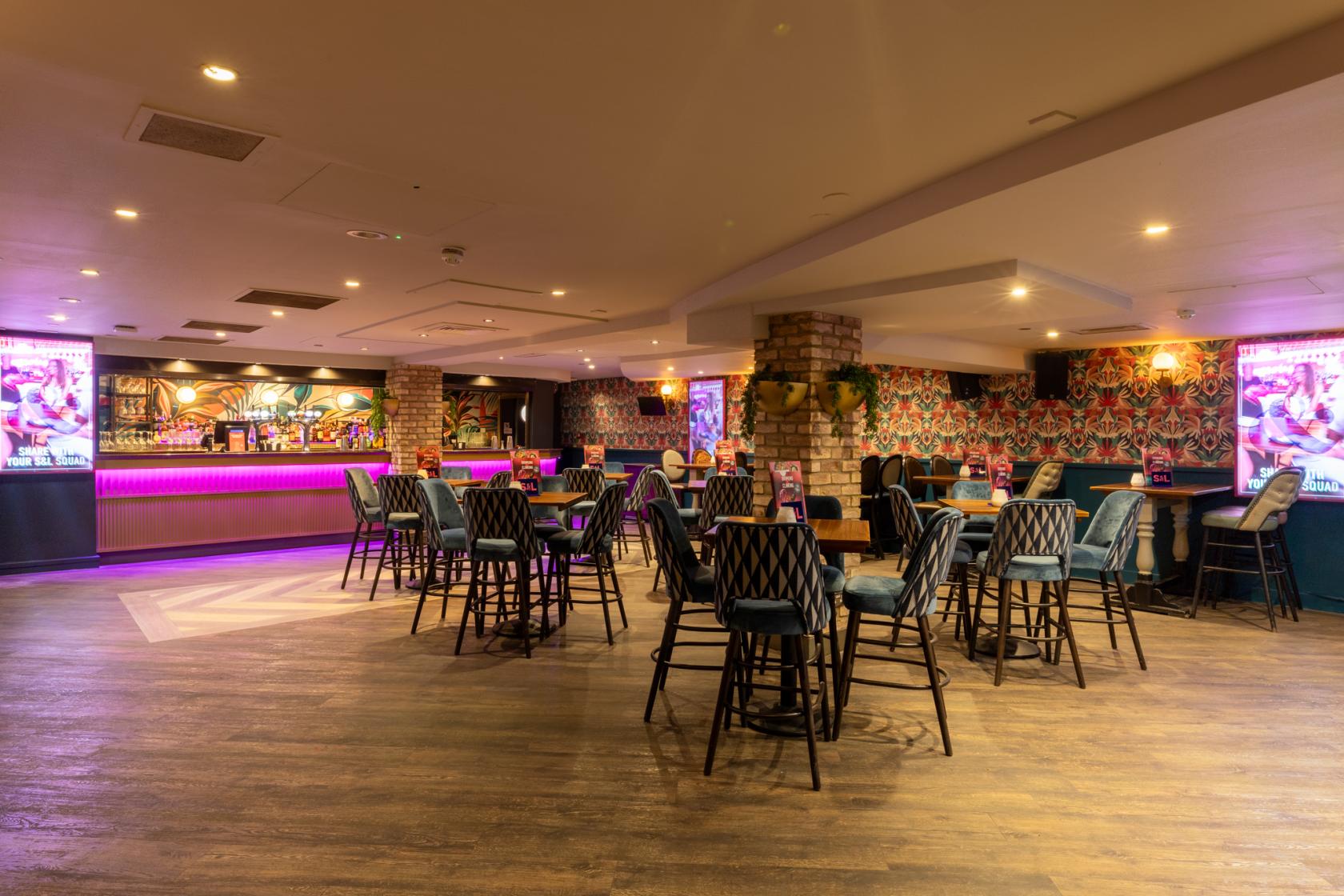 A function room in a cocktail bar with high tables, patterned chairs, neon-lit bar, and colorful wallpaper at Slug & Lettuce Waterloo.