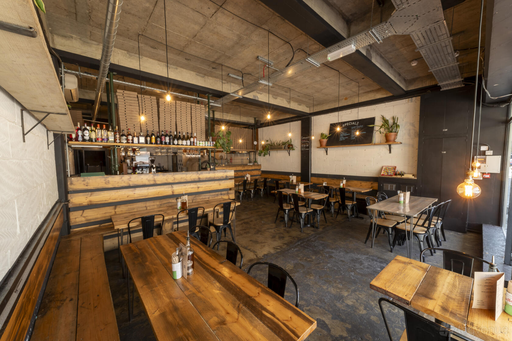 A restaurant with wooden communal tables, hanging pendant lights, and open shelving at Mamma Dough Sydenham.