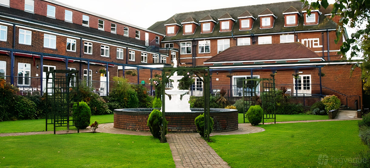 A hotel courtyard with a brick walkway, white tiered fountain, manicured lawn, and trimmed bushes at The Thurrock Hotel.