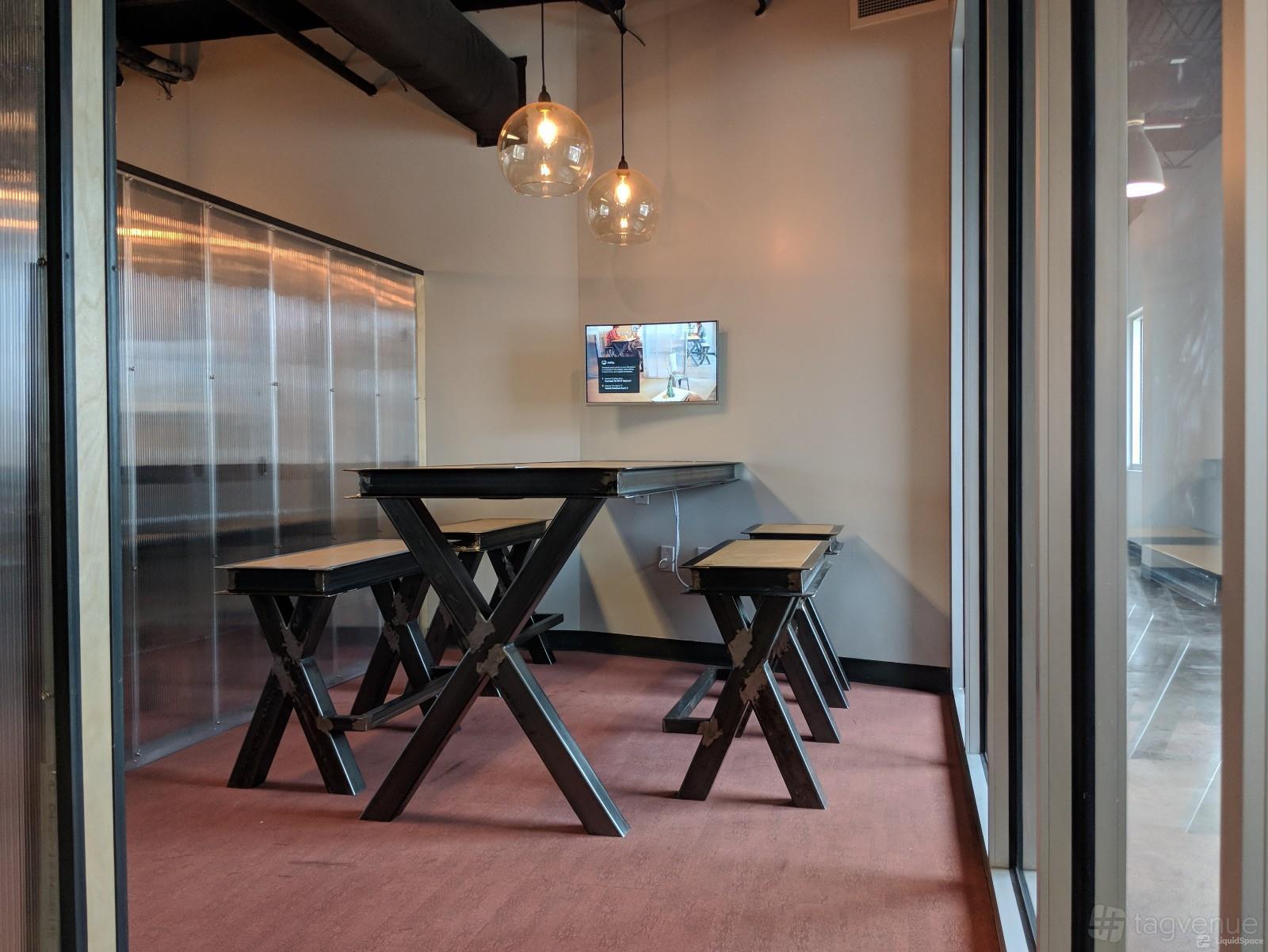 A coworking meeting room with high-top tables, benches, pendant lighting, and a wall-mounted screen at Vessel Coworking.